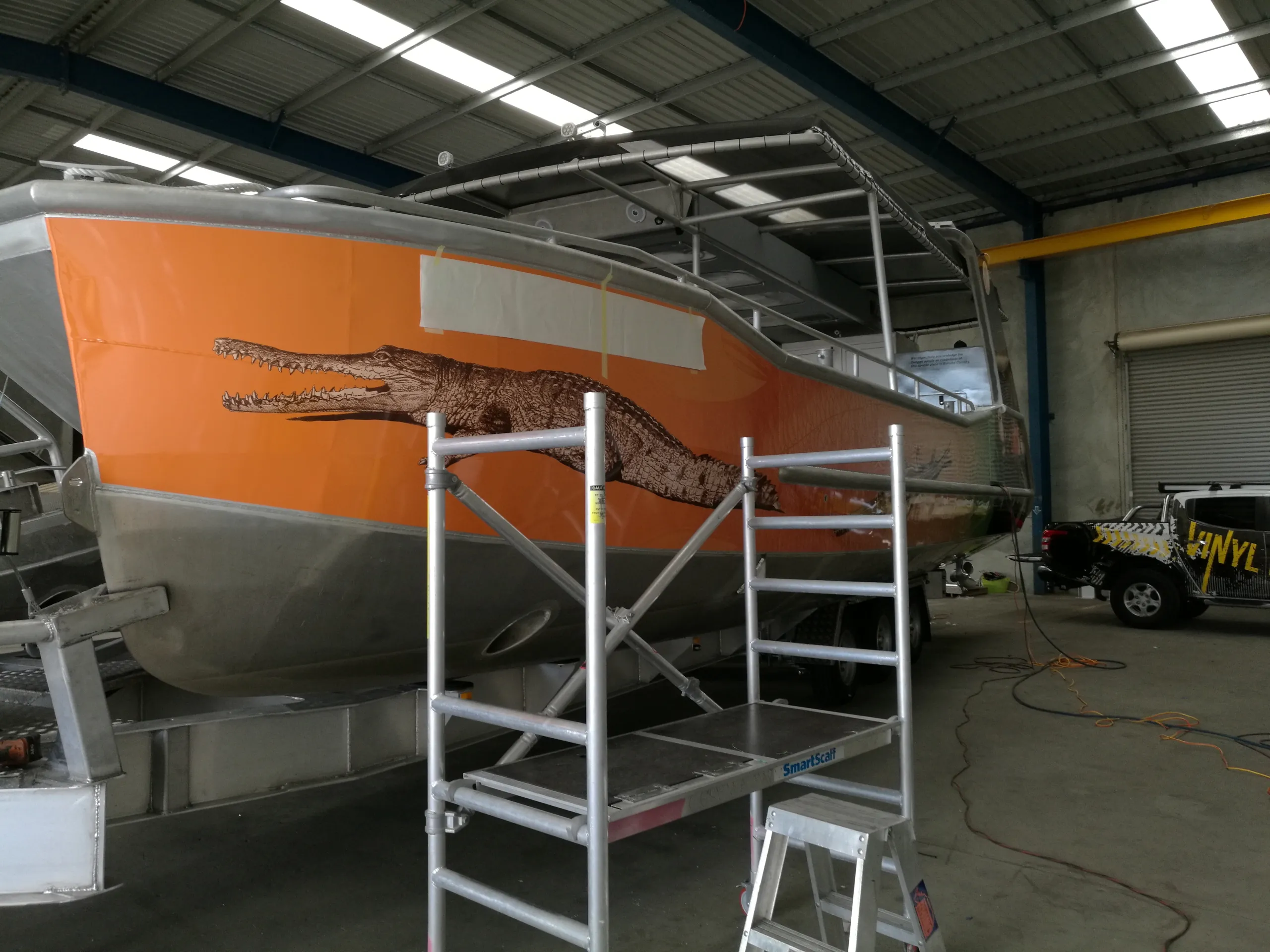 Orange crocodile graphics on a boat bow during installation, with masking tape and scaffolding