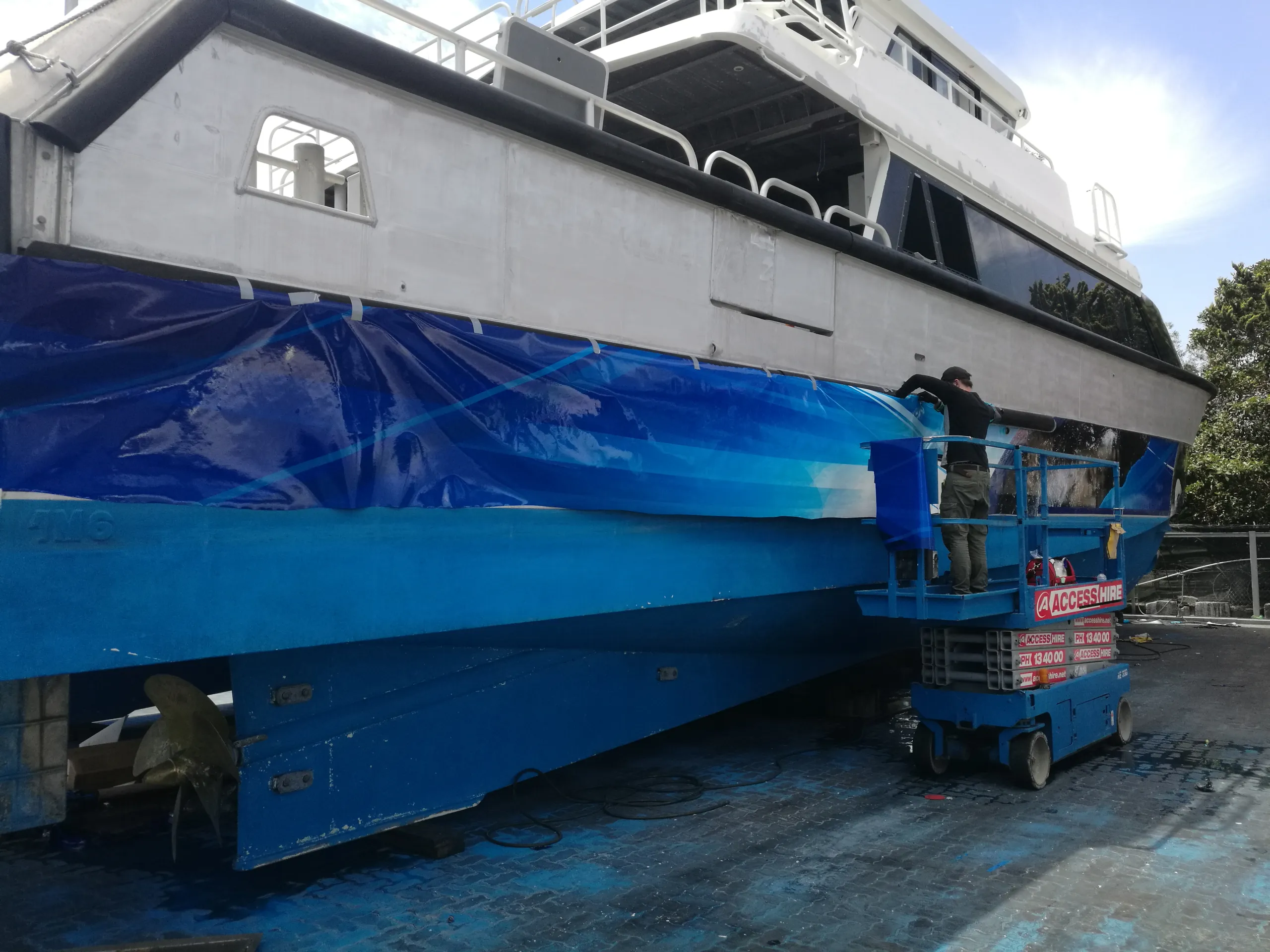 Installer fitting blue graphics to the Naturaliste Charters catamaran from a scissor lift outdoors