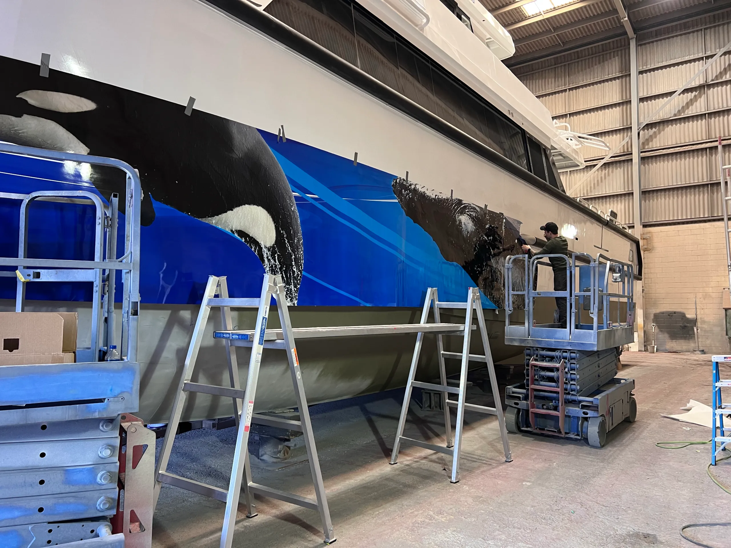 Installer aligning whale graphics along the Naturaliste Charters catamaran hull from a scissor lift