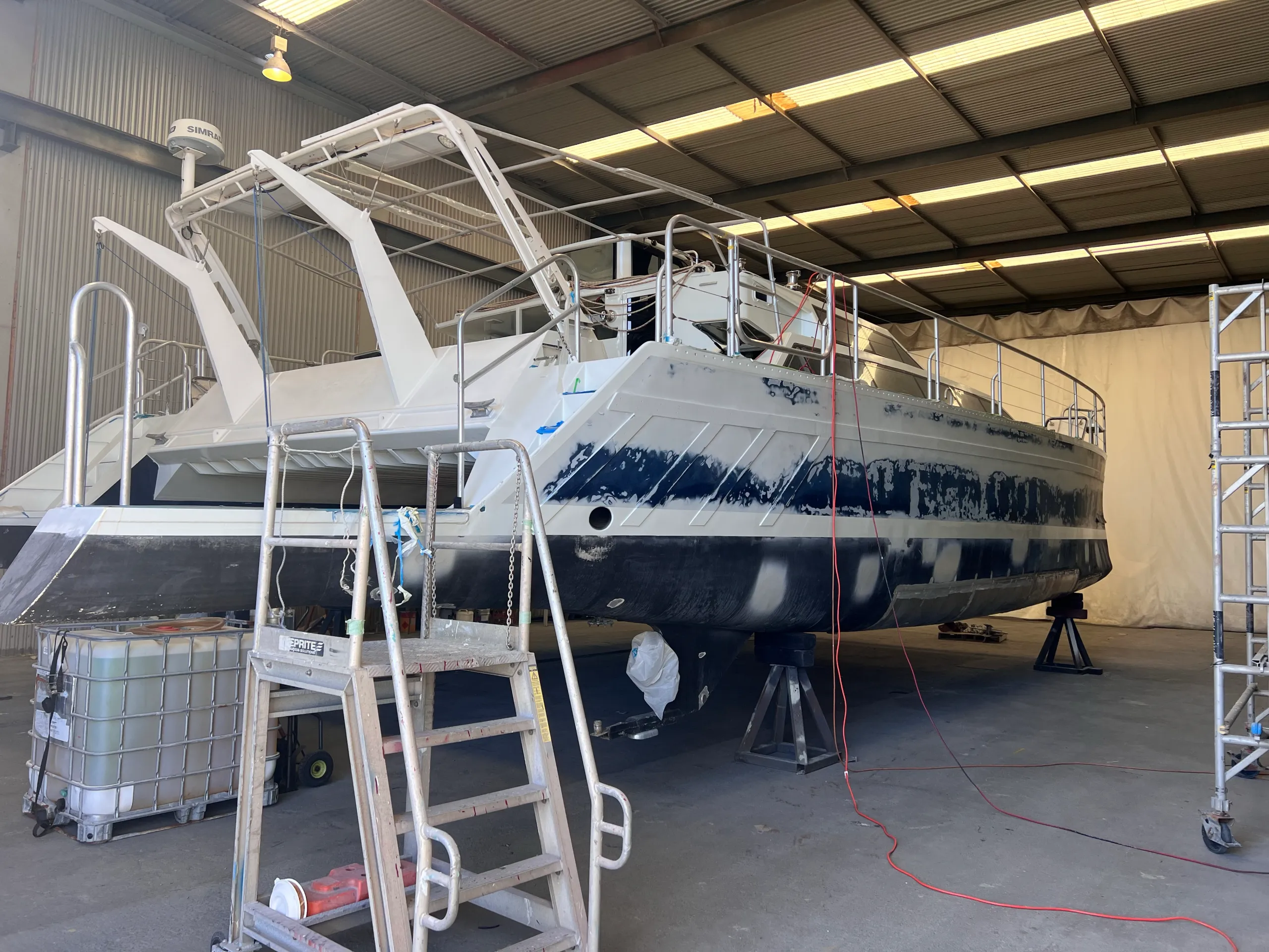 Catamaran stern on stands inside a shed, with patchy hull surfaces during preparation for new graphics