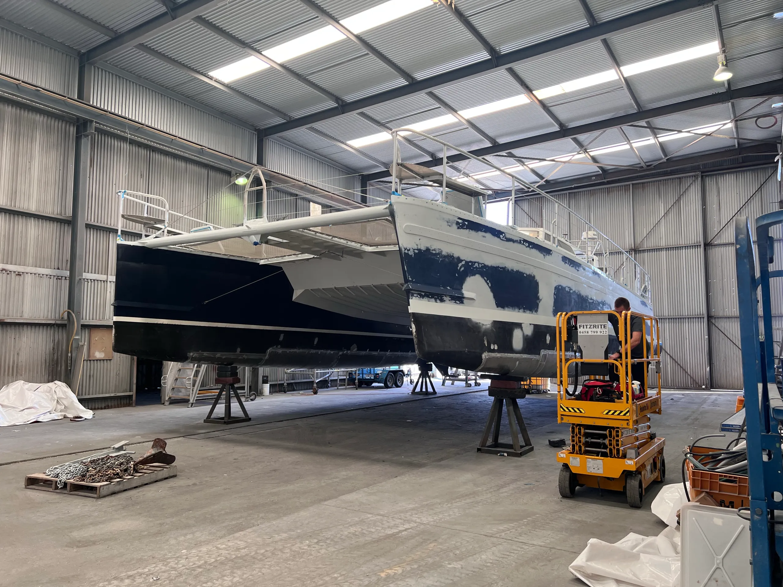 Catamaran bows during hull preparation, with a worker on an access lift beside the boat