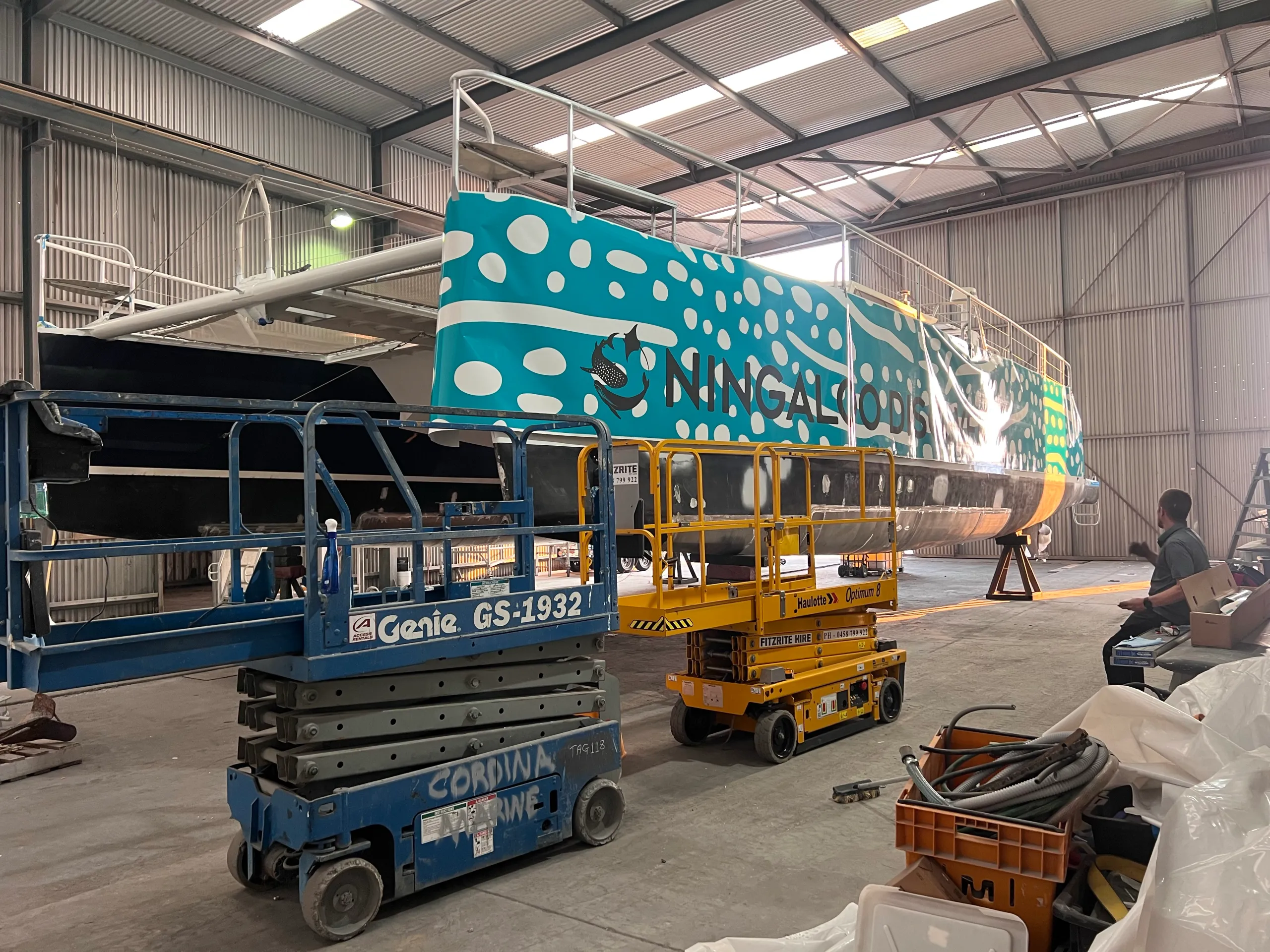 Turquoise and white Ningaloo Discovery graphics being fitted to a catamaran hull inside a shed
