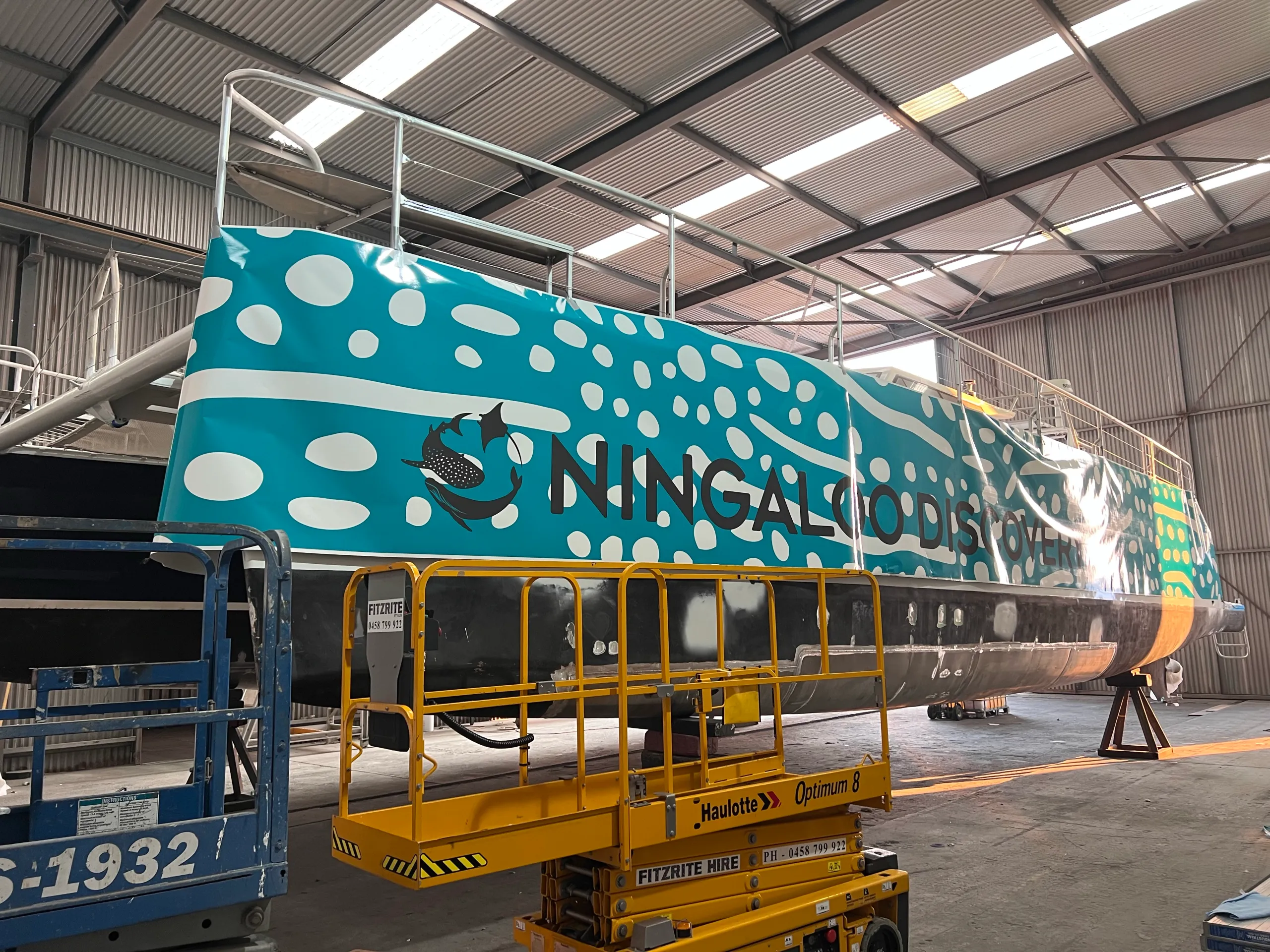 Ningaloo Discovery lettering across turquoise whale-shark-pattern graphics on a catamaran hull during installation
