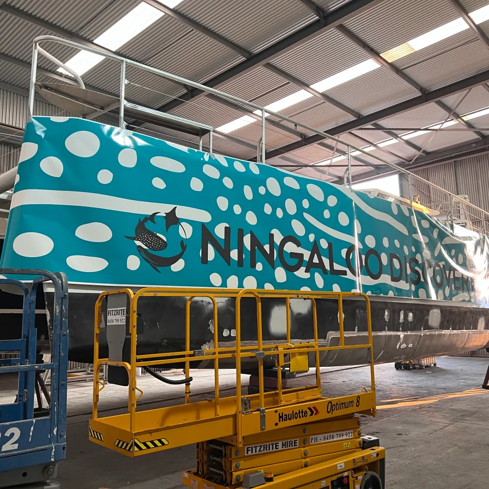 Ningaloo Discovery lettering across turquoise whale-shark-pattern graphics on a catamaran hull during installation