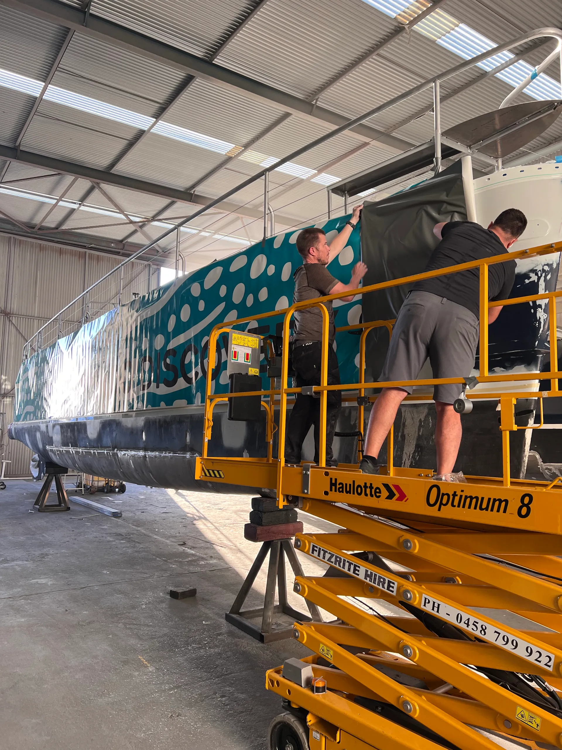 Installers on an access platform fitting turquoise printed film along the catamaran hull