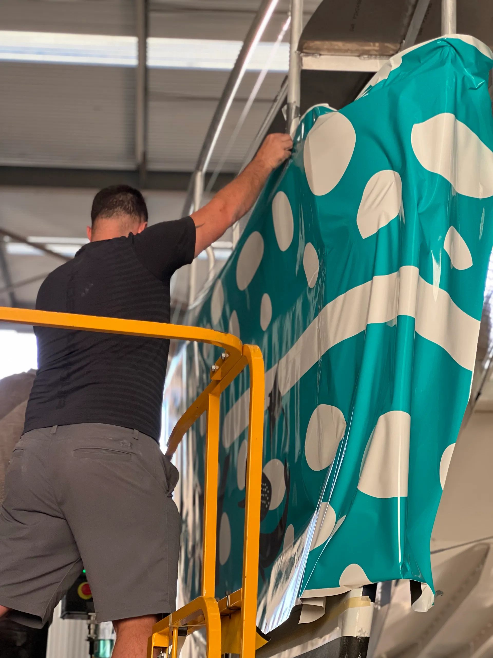 Back-facing installer holding turquoise and white film against the catamaran hull from an access platform