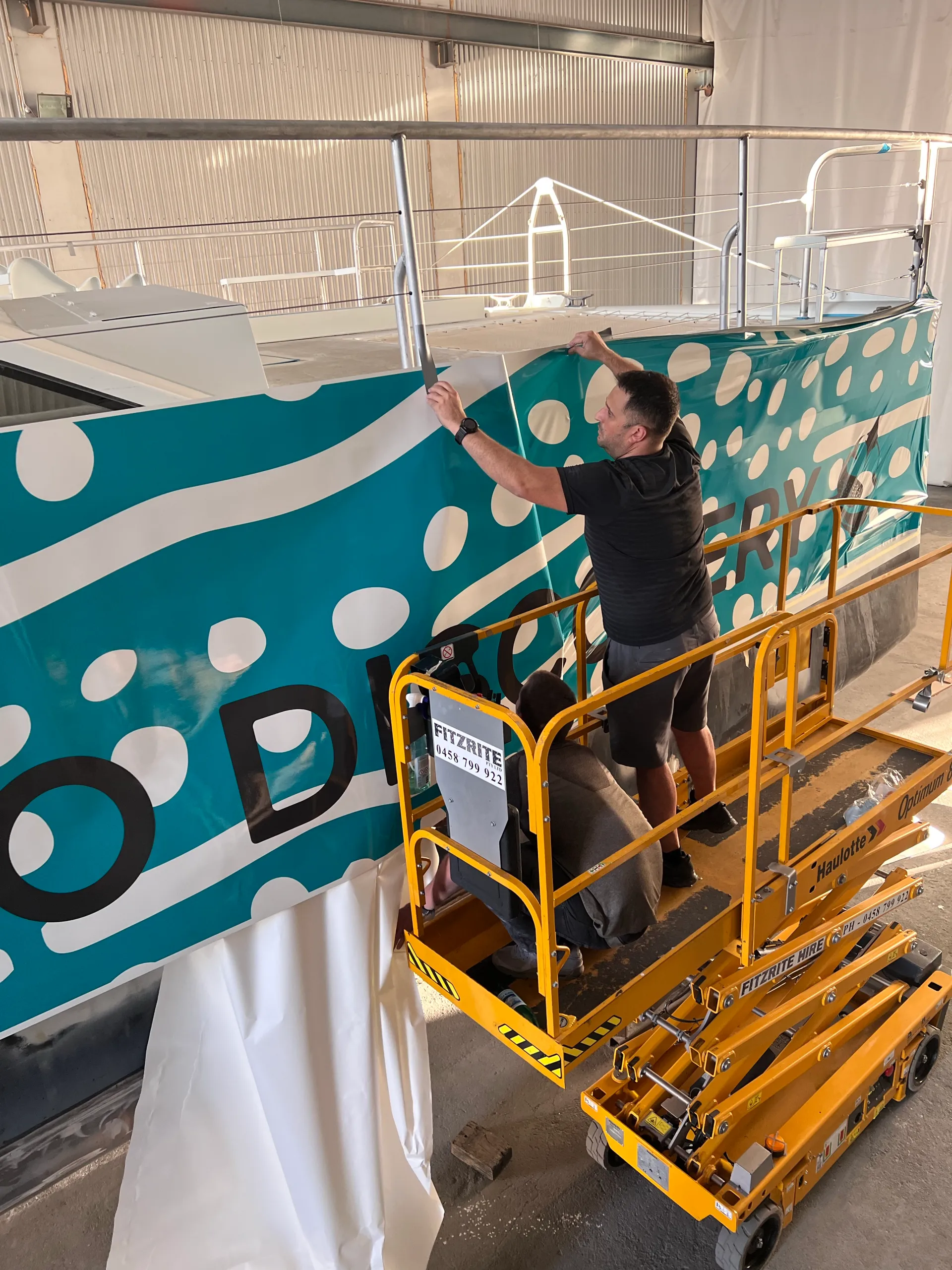 Installer smoothing Ningaloo Discovery graphics along the catamaran hull from a raised platform