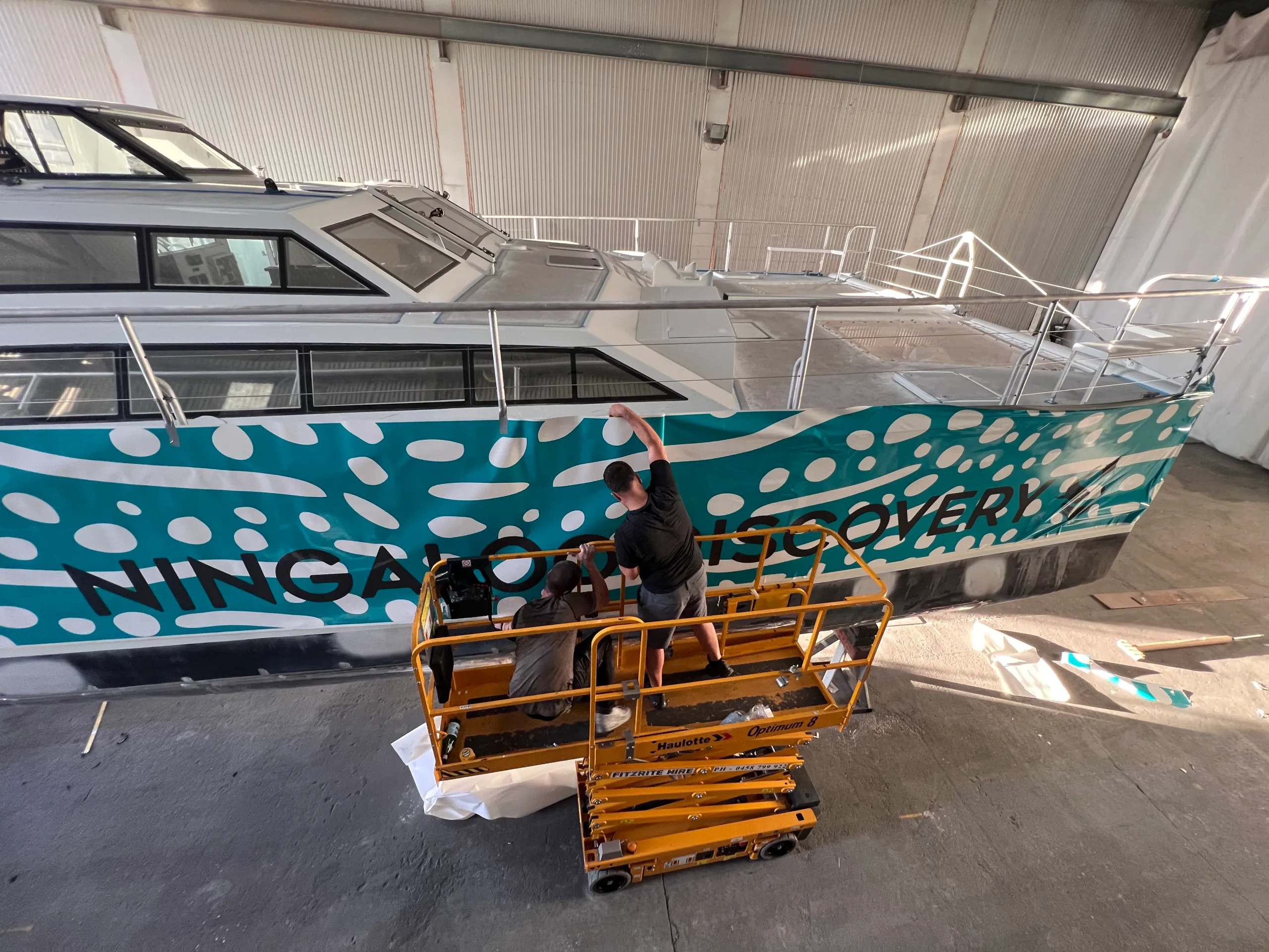 Elevated view of installers applying turquoise and white Ningaloo Discovery graphics to the boat hull