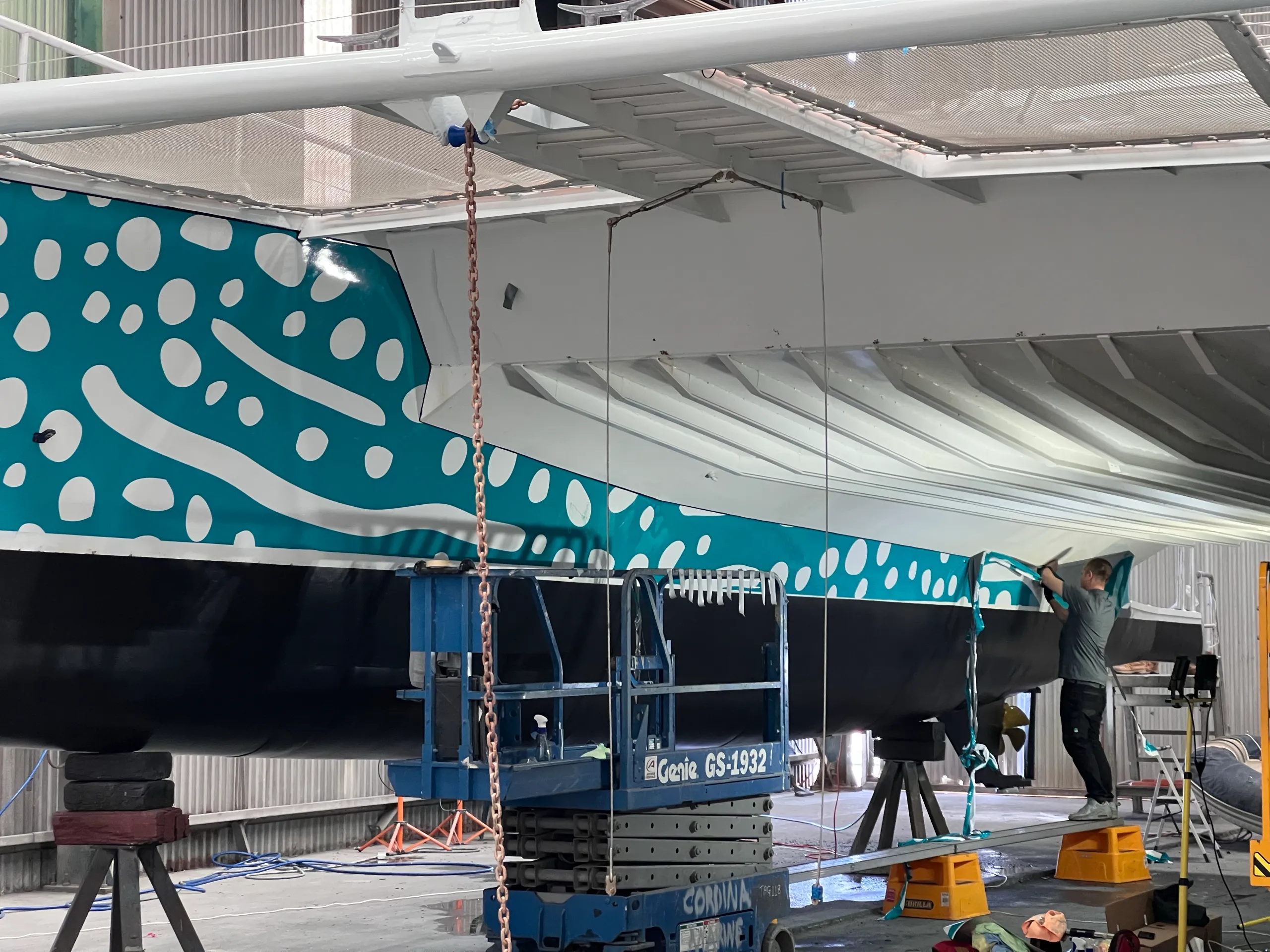 Installer fitting turquoise spotted graphics along the inner hull beneath the catamaran's forward deck