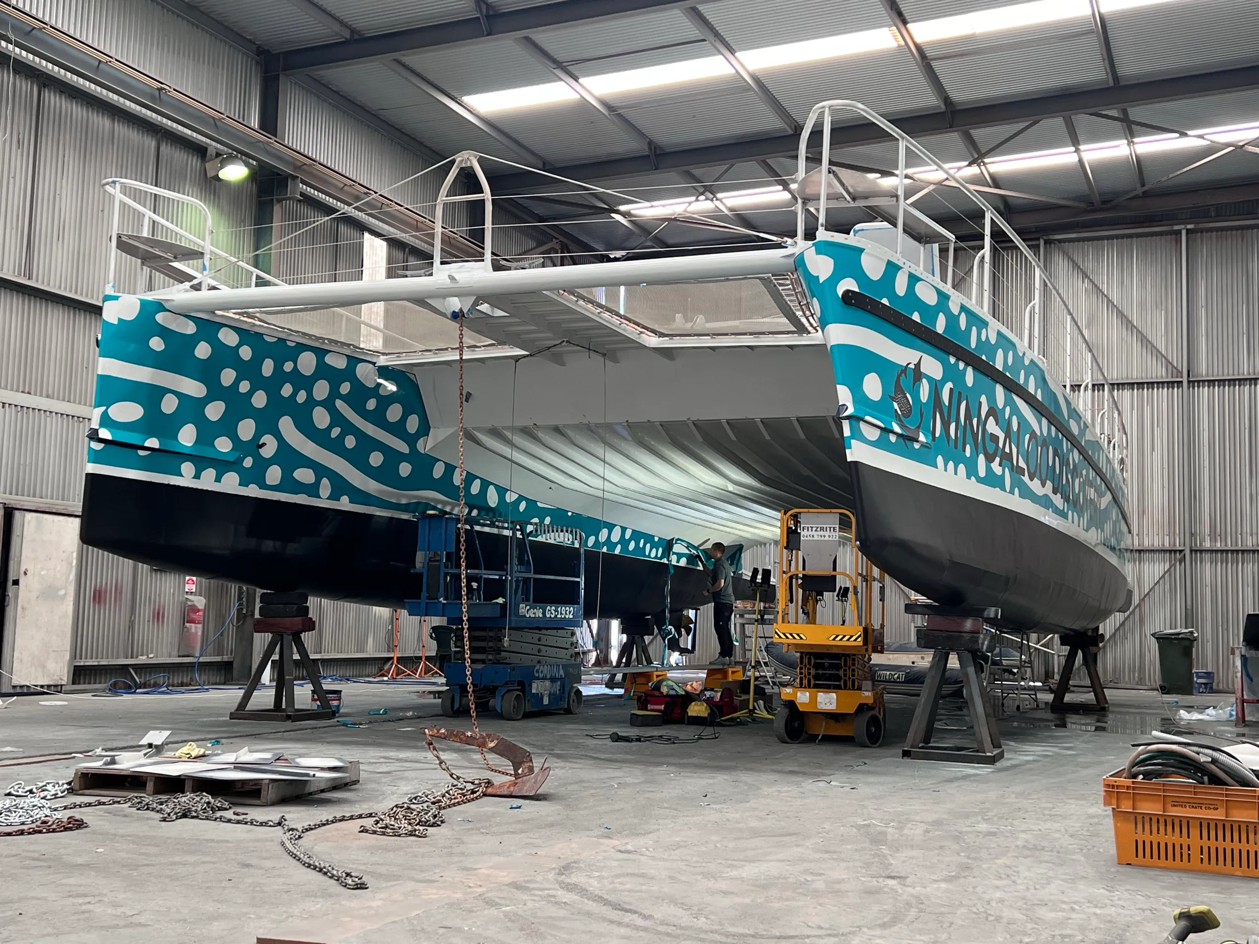 Catamaran bows on stands with turquoise spotted graphics, as installers work beneath the forward deck