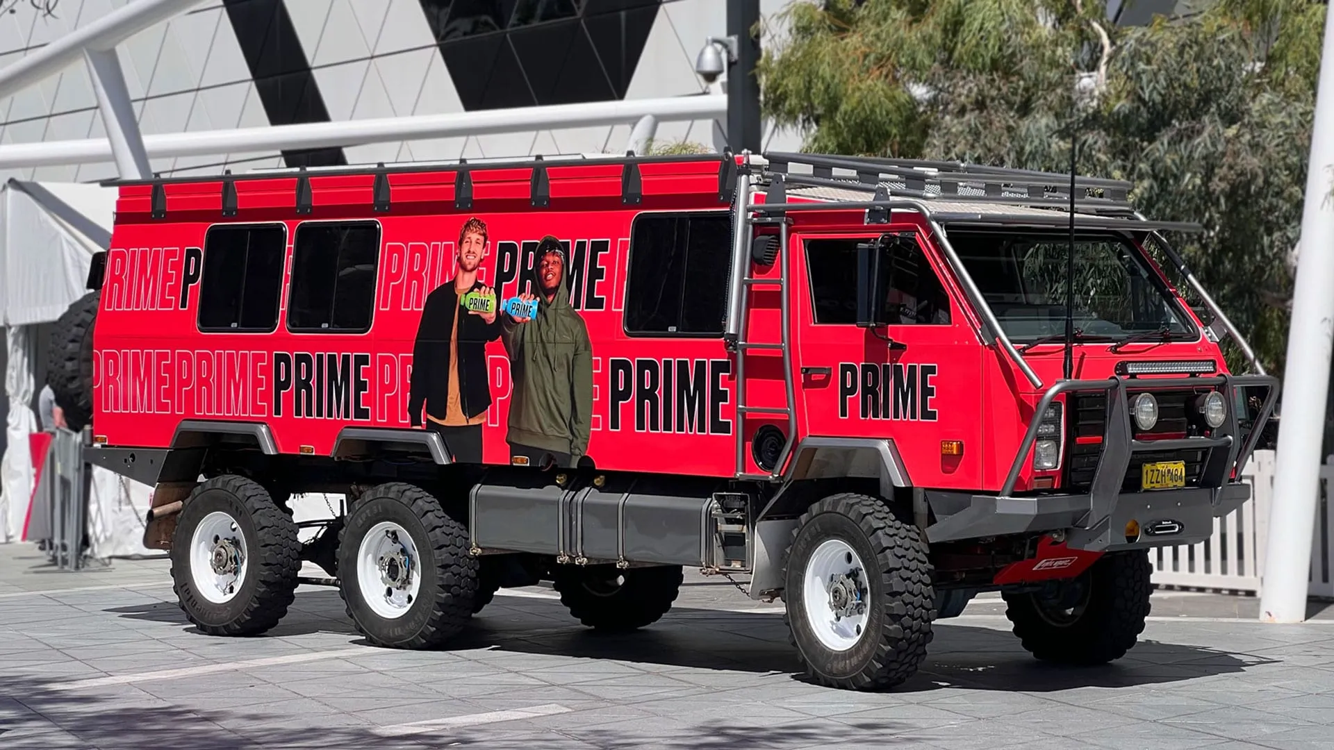 OKA 6x6 expedition truck in a full printed red Prime Hydration wrap, parked outside a stadium
