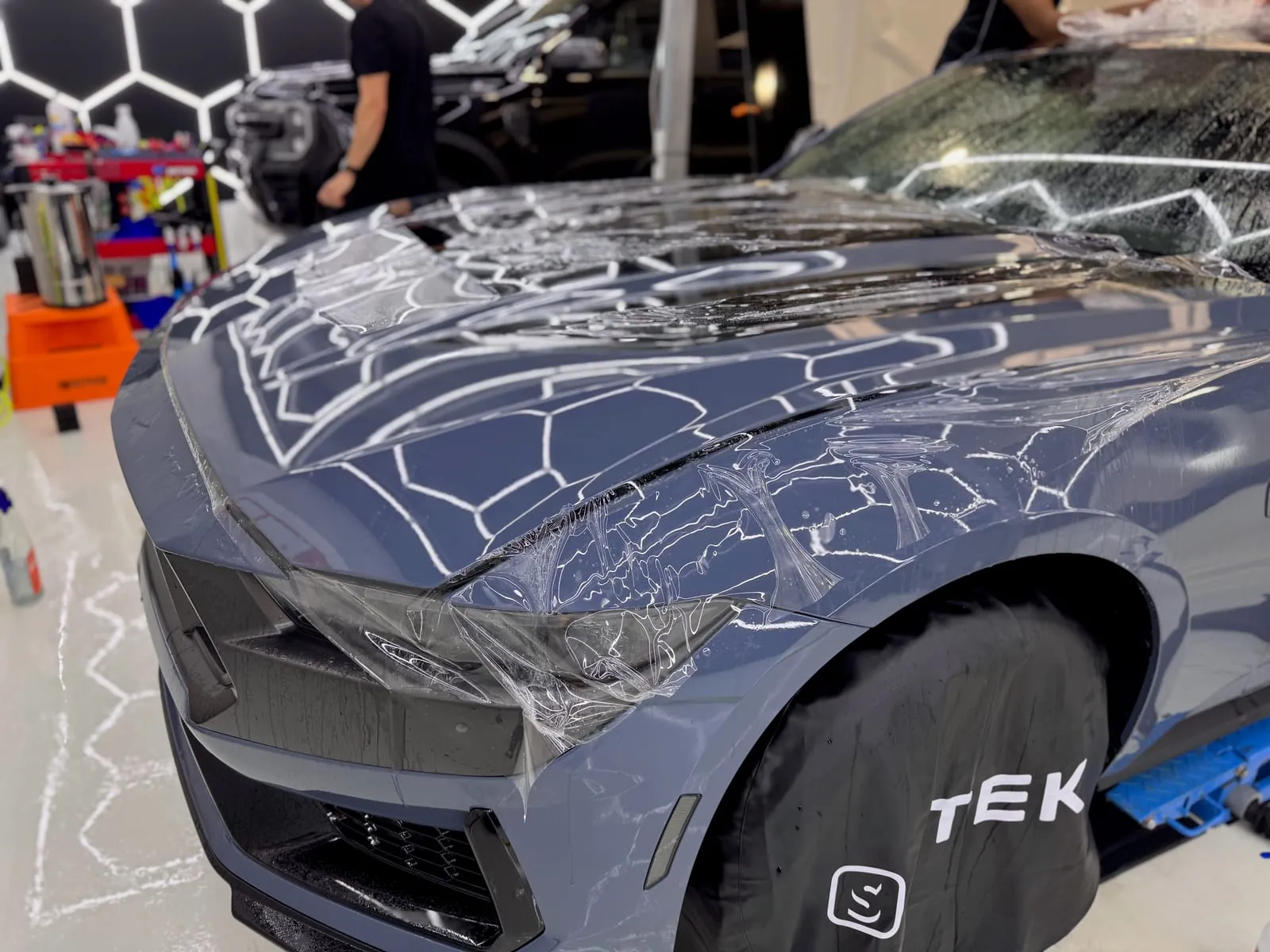 Clear paint protection film being laid over the bonnet of a grey Mustang under the hexagon lighting