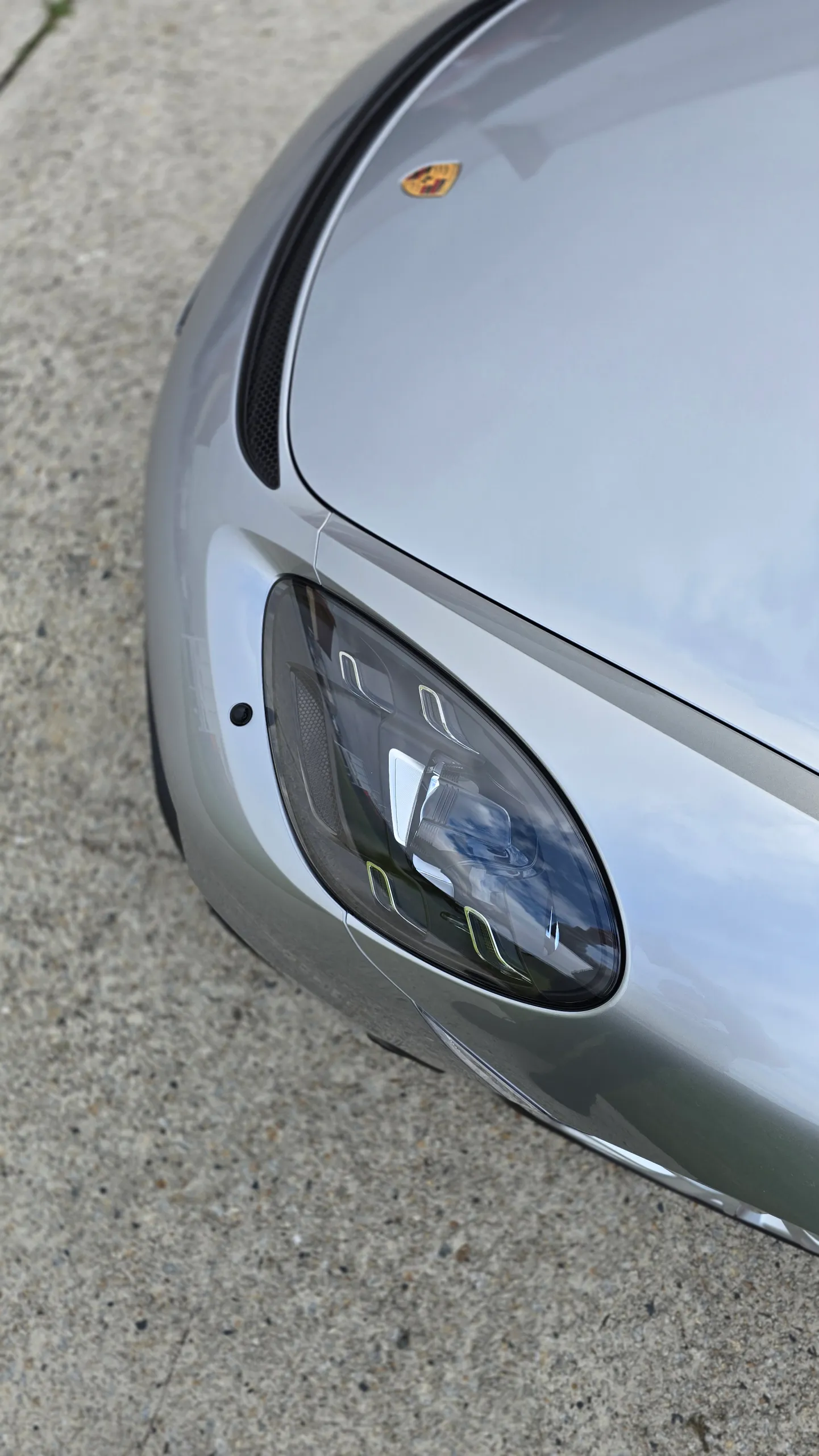 Porsche crest and headlight beside the silver 718 Spyder bonnet