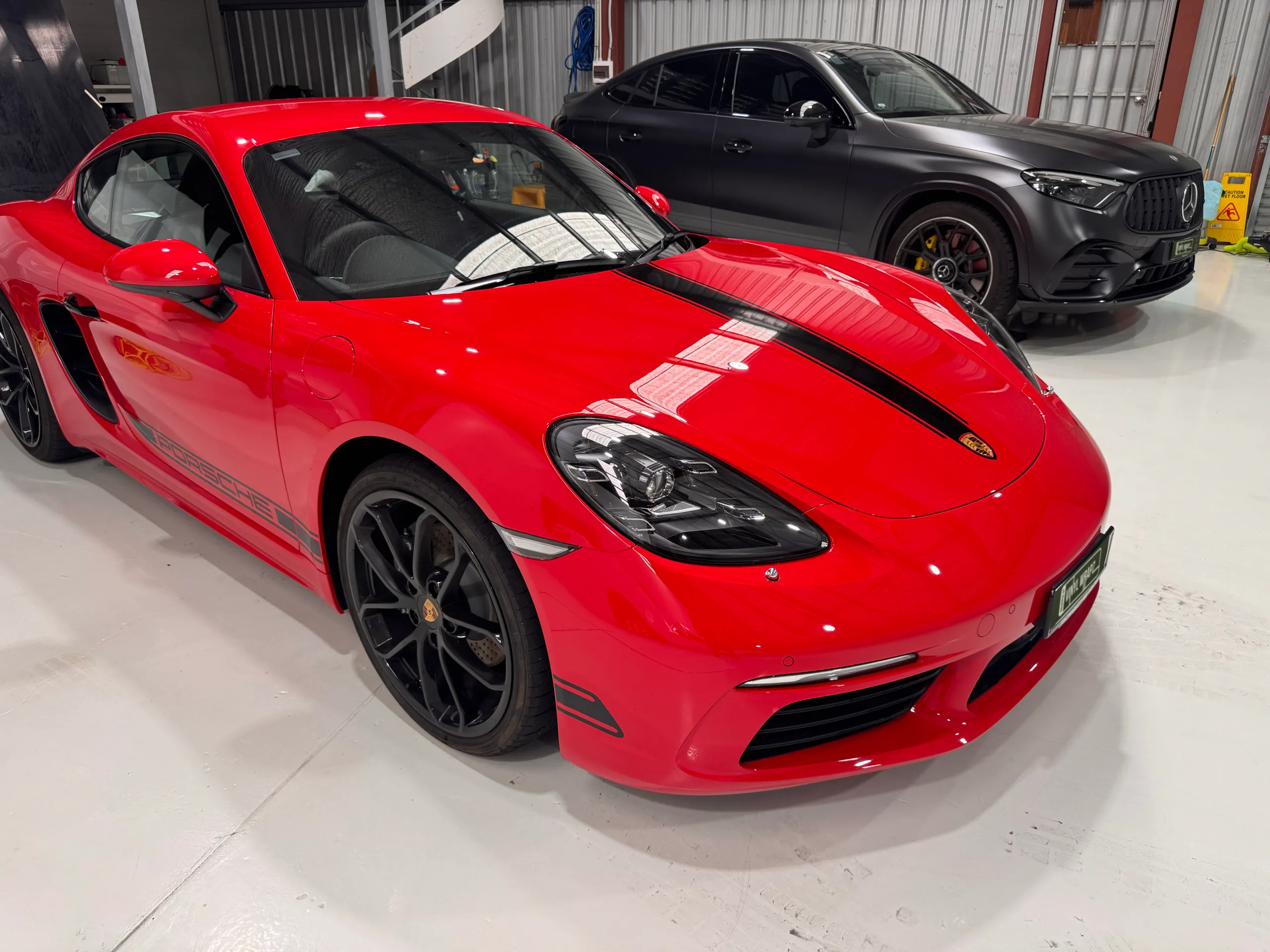 Gloss red Porsche Cayman with black bonnet and side stripes, viewed from the front quarter in the workshop