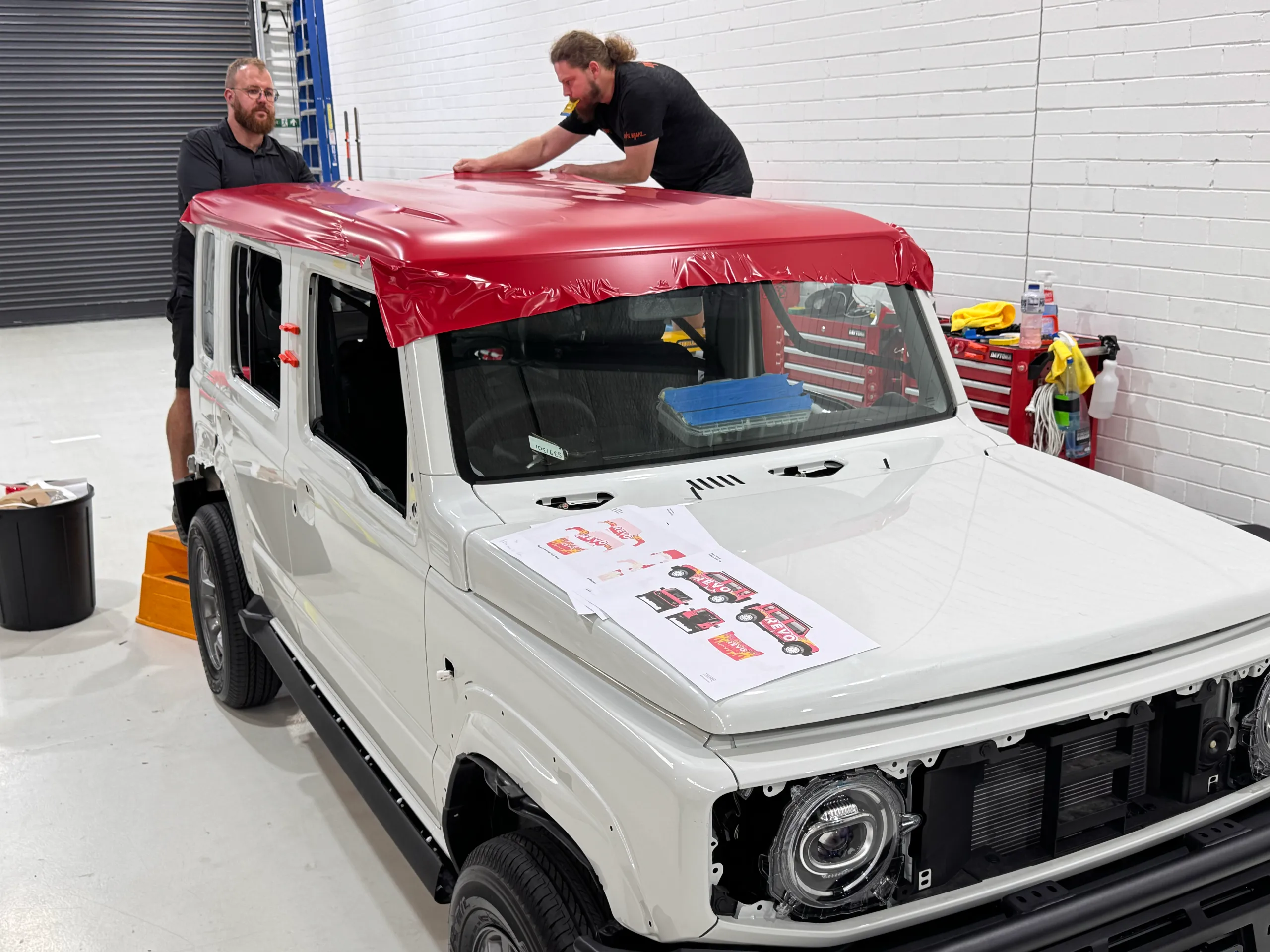 Two installers fitting red film to the white Suzuki Jimny roof