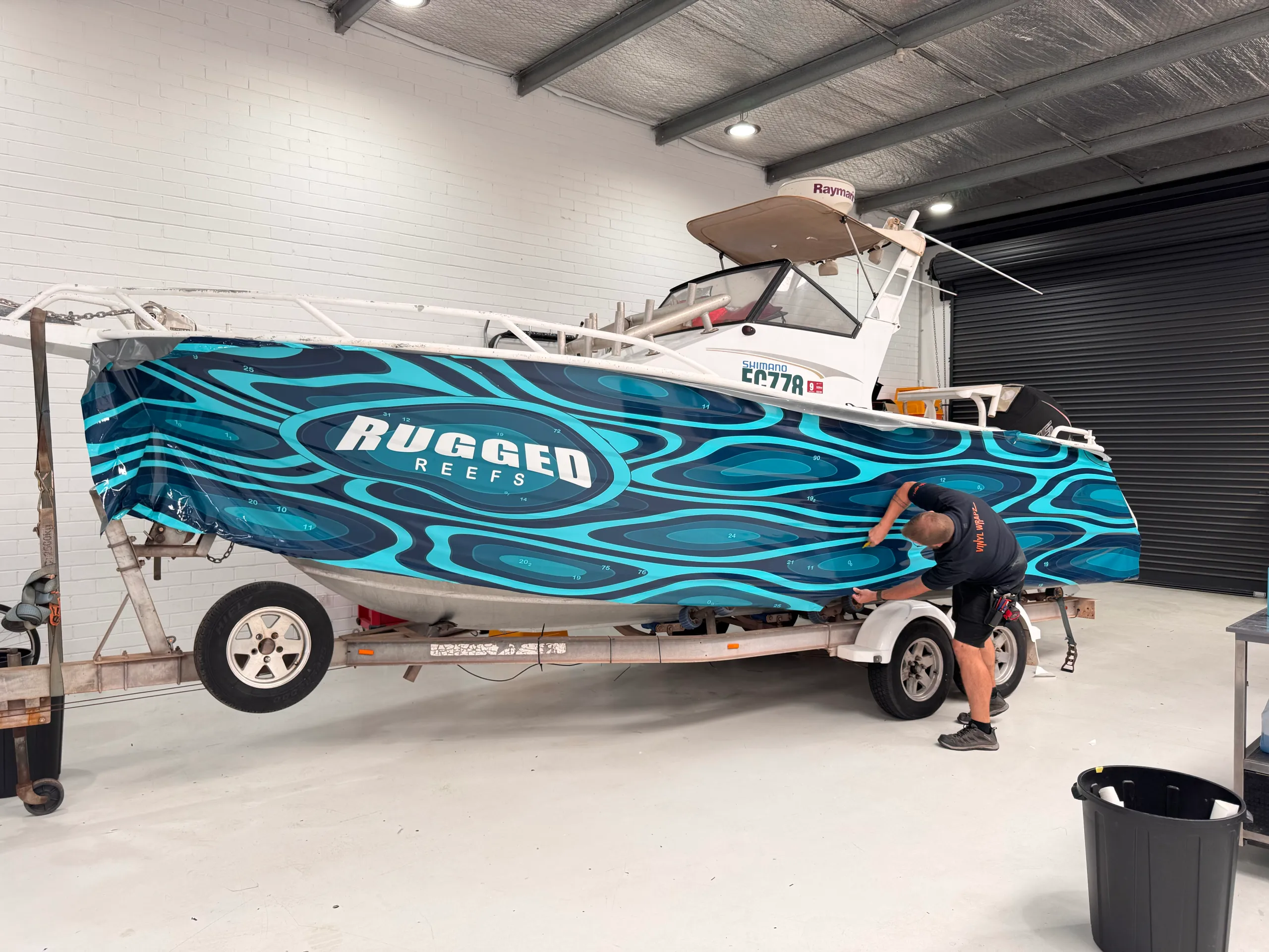 Teal topographic film being fitted along the hull, with an installer working near the stern