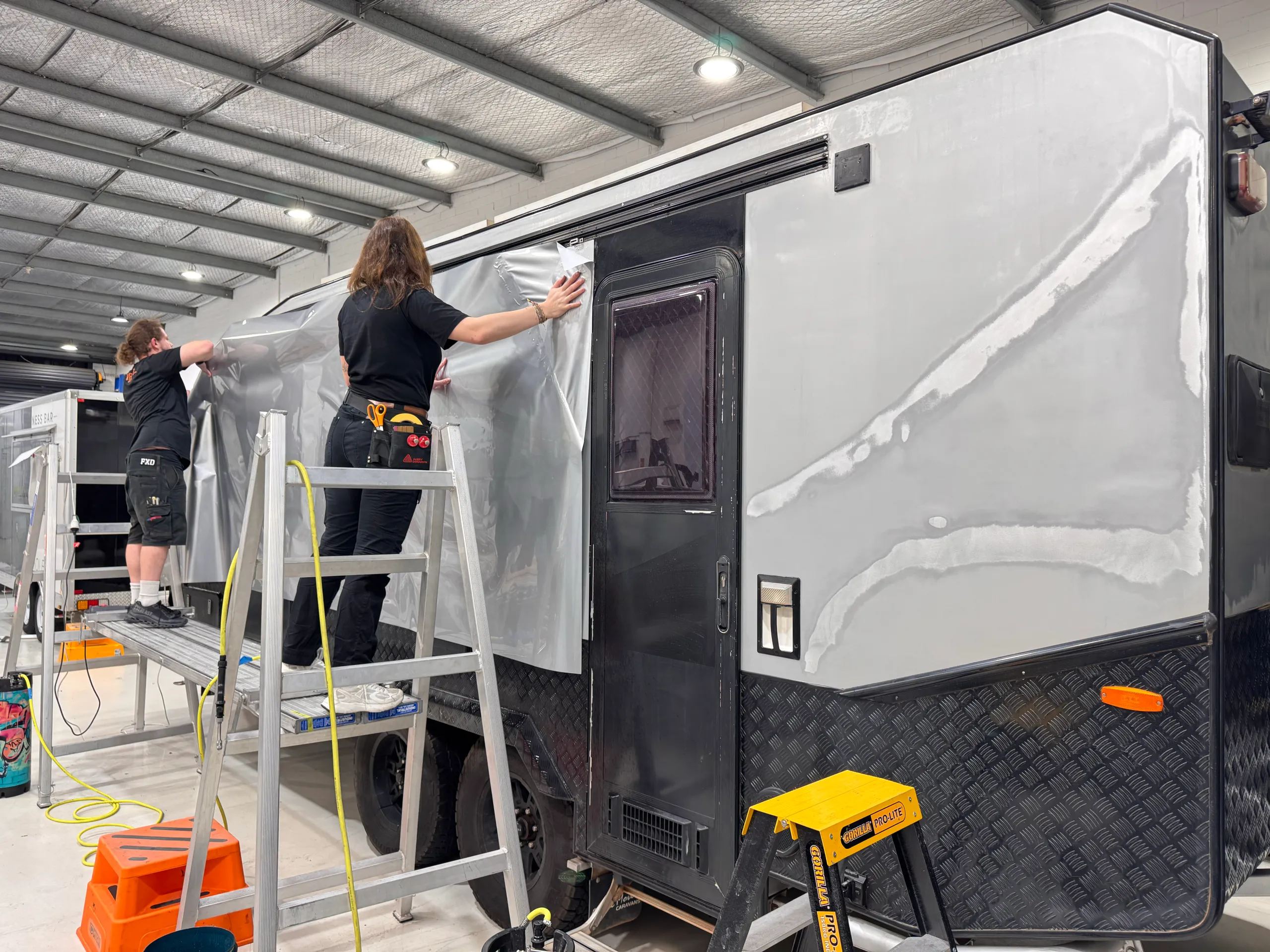 Installers seen from behind fitting silver film along a prepared caravan side panel