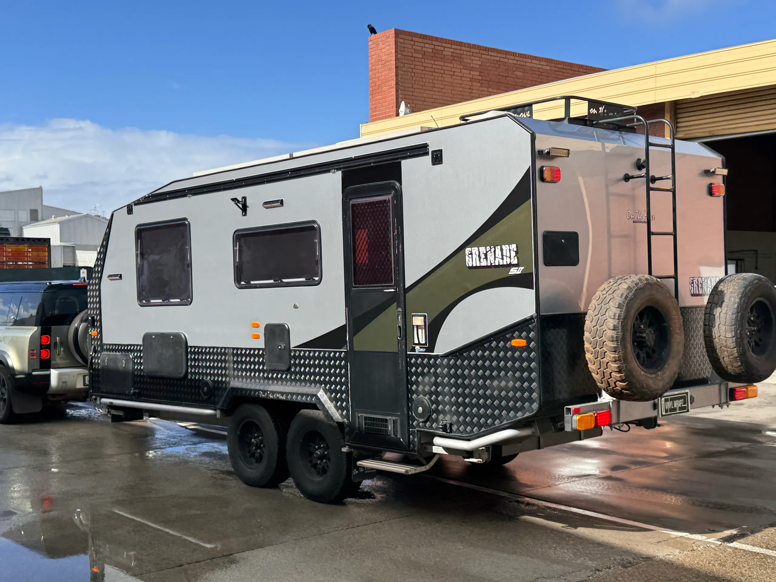 Rear-quarter view of the silver caravan with olive green graphics and two spare wheels outdoors