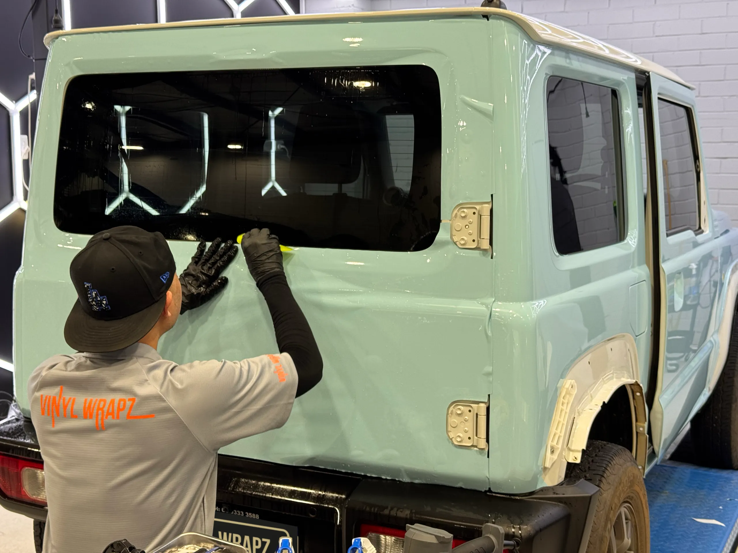 Back-facing installer smoothing light blue film below the Suzuki Jimny rear window