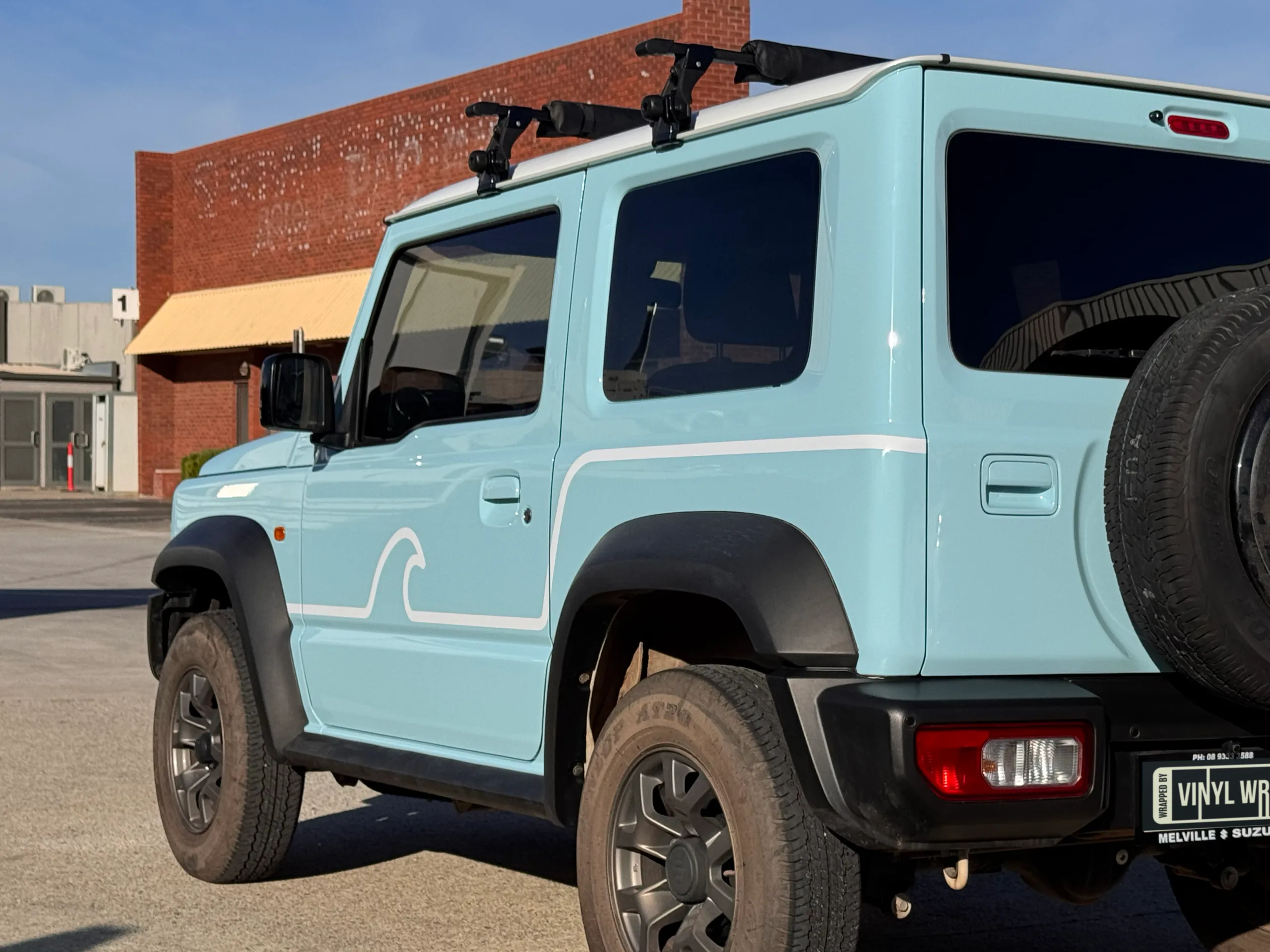 Rear-quarter view of the light blue Suzuki Jimny with a white wave graphic on the door
