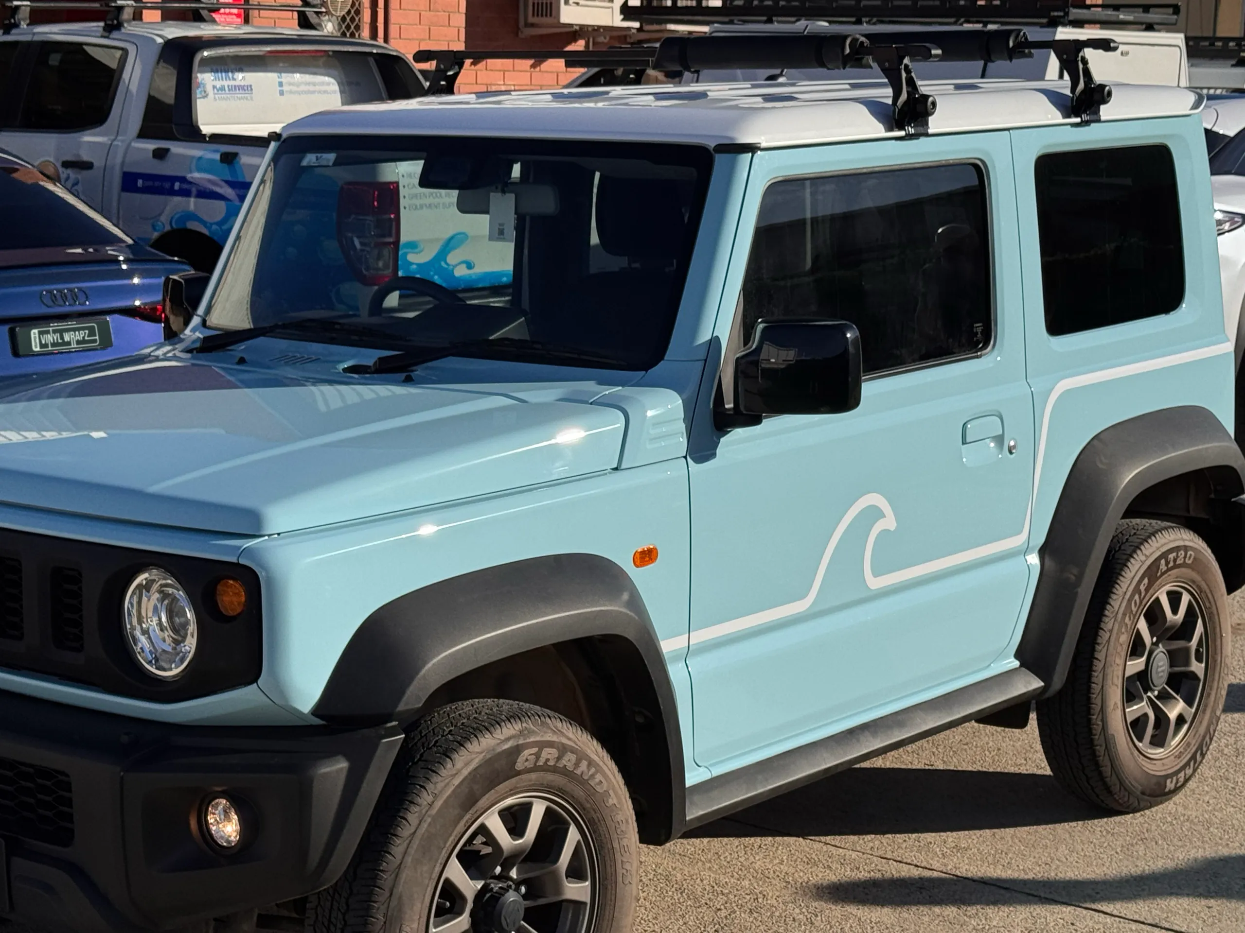 Light blue Suzuki Jimny with a white wave door graphic and black roof bars outdoors