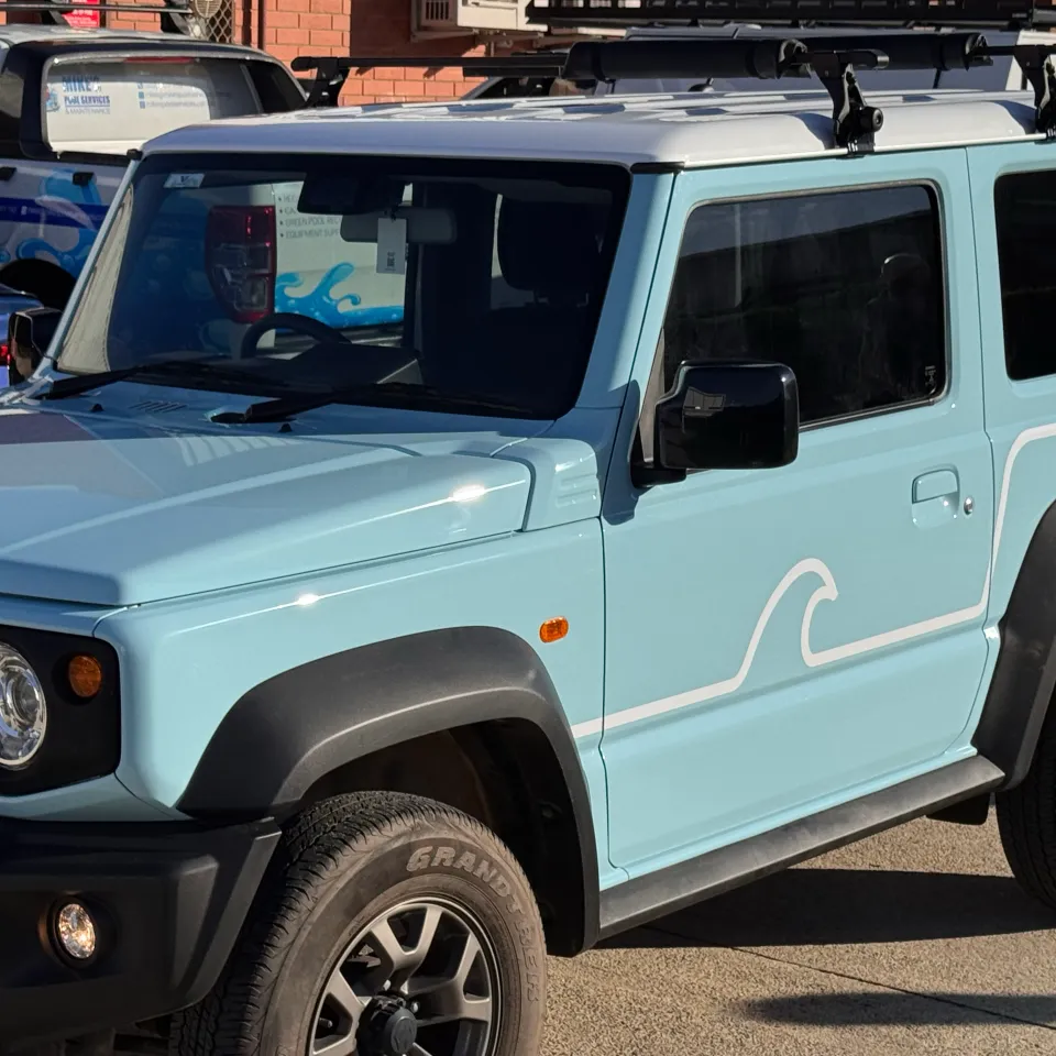 Light blue Suzuki Jimny with a white wave door graphic and black roof bars outdoors