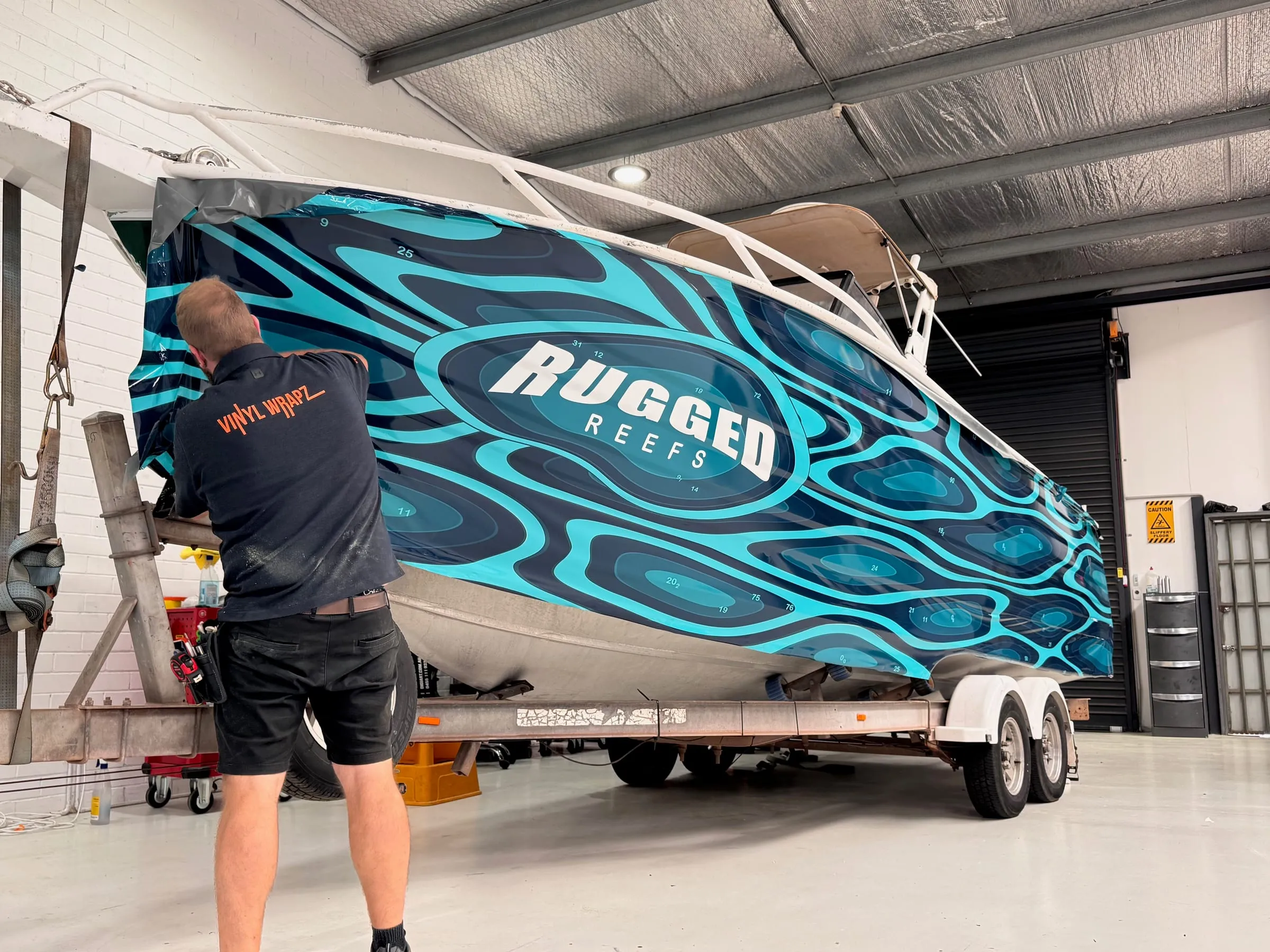 Vinyl Wrapz installer applying a printed wave-pattern wrap to a fishing boat bow in the Osborne Park workshop