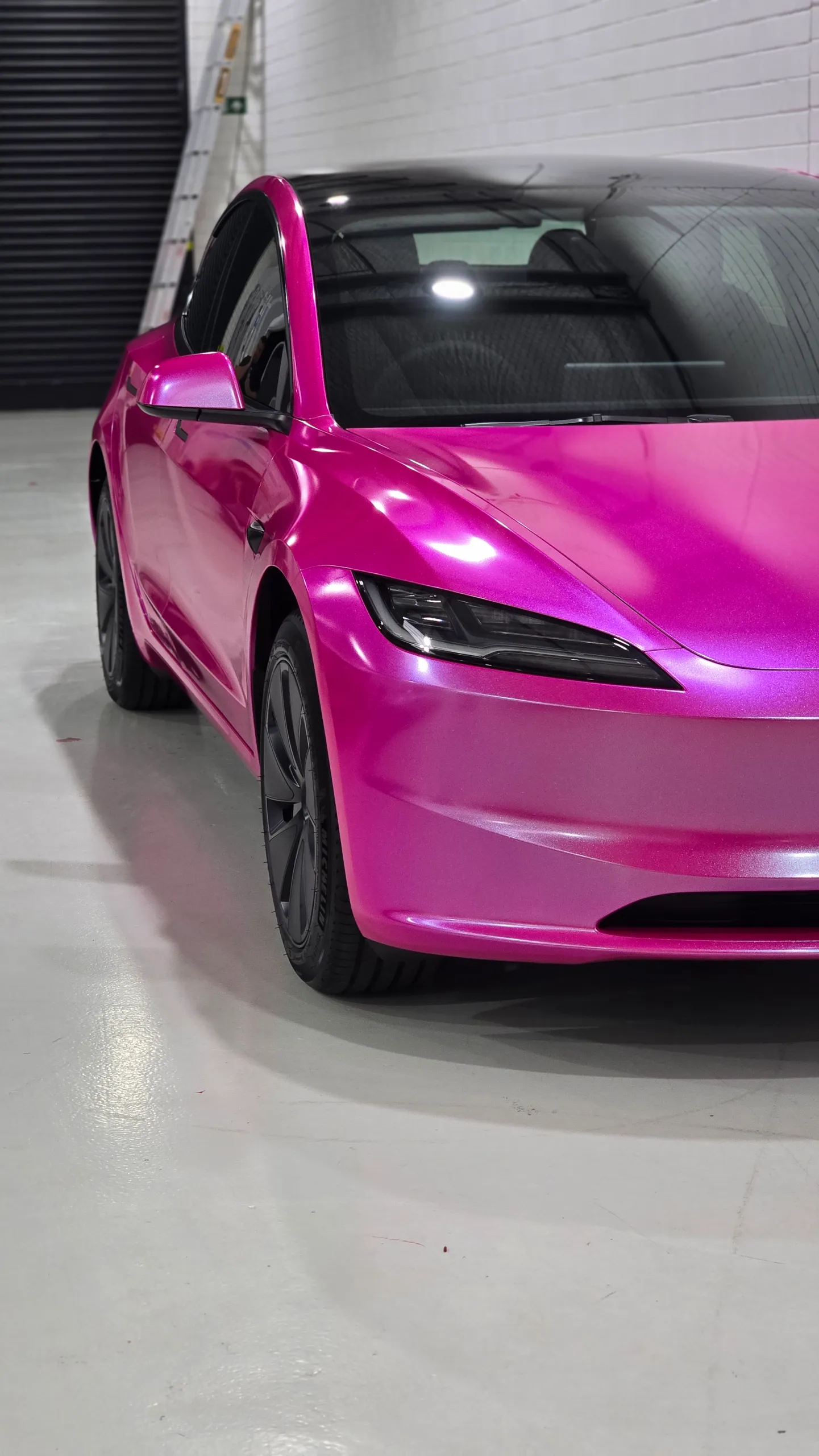Pink Tesla Model 3 viewed from a cropped front angle beside a white brick wall