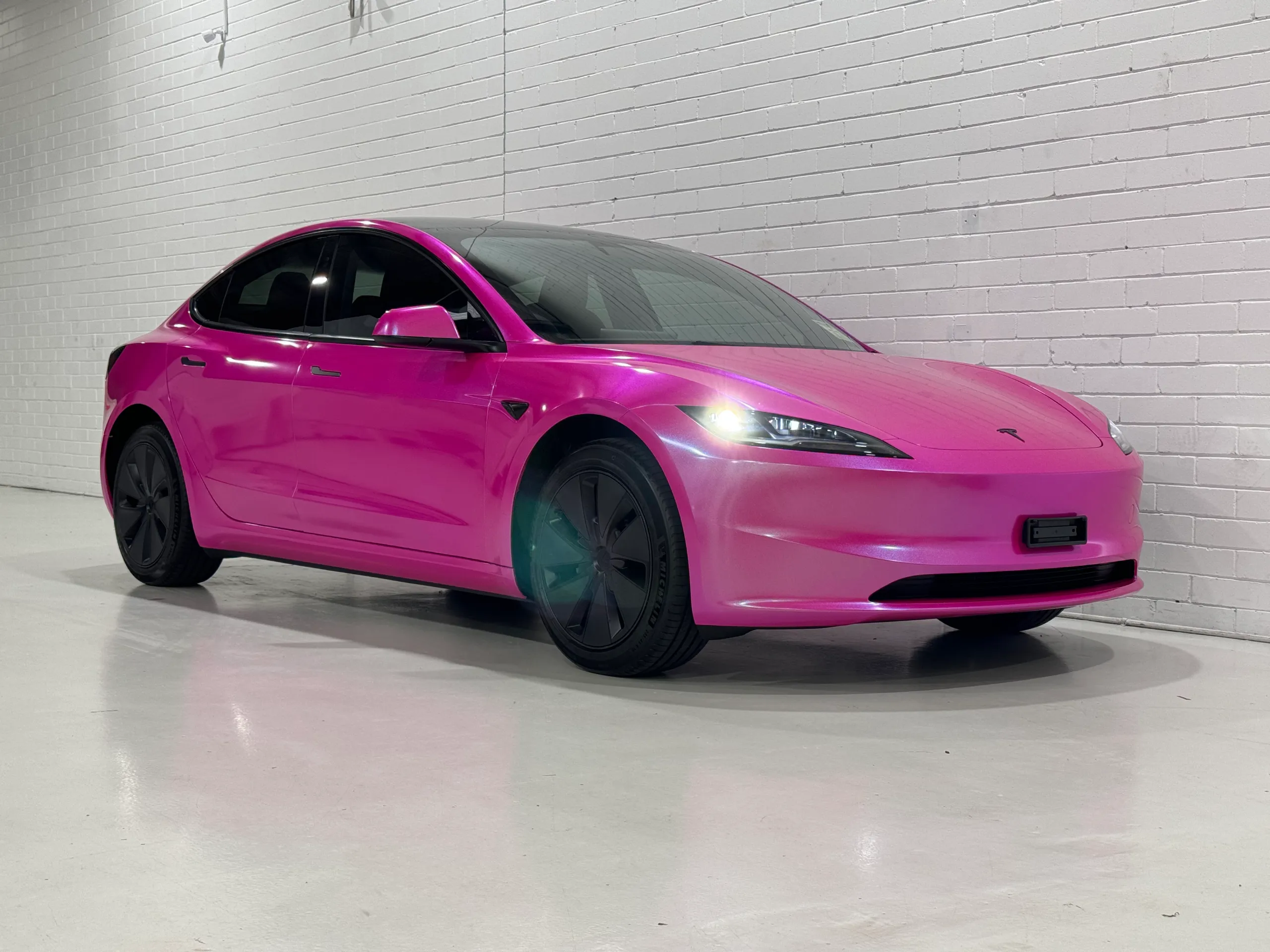 Gloss pink Tesla Model 3 viewed from the front quarter with its headlights on