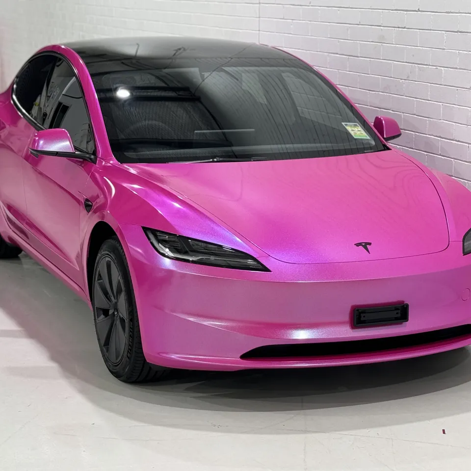 Elevated front-quarter view of the pink Tesla Model 3 beside a white workshop wall