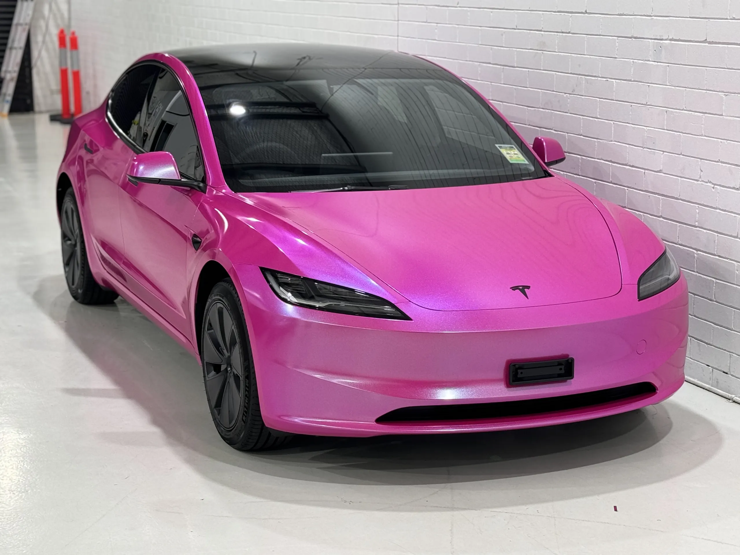 Elevated front-quarter view of the pink Tesla Model 3 beside a white workshop wall