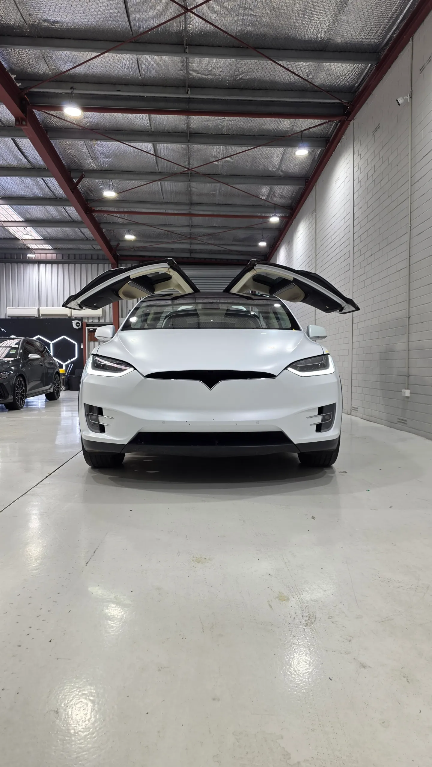White Tesla Model X viewed head-on with both falcon-wing doors raised in the workshop