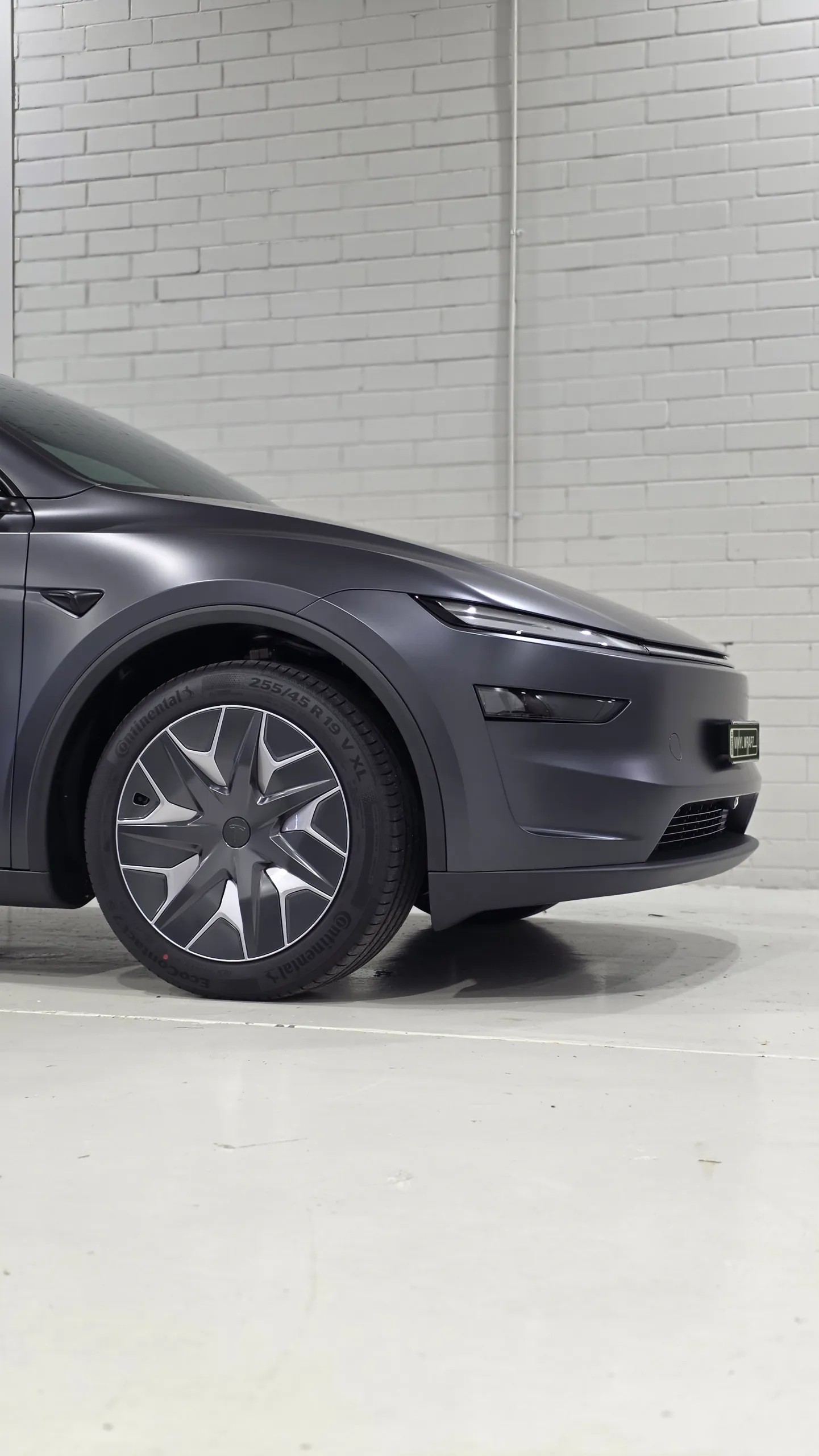 Tesla Model Y front wheel and matte dark grey front bodywork against a white workshop wall