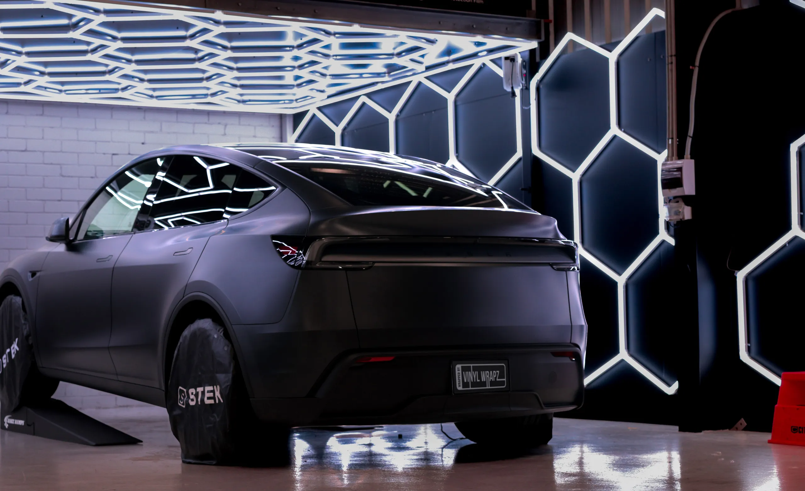 Satin dark Tesla Model Y beneath hexagonal workshop lights with protective wheel covers