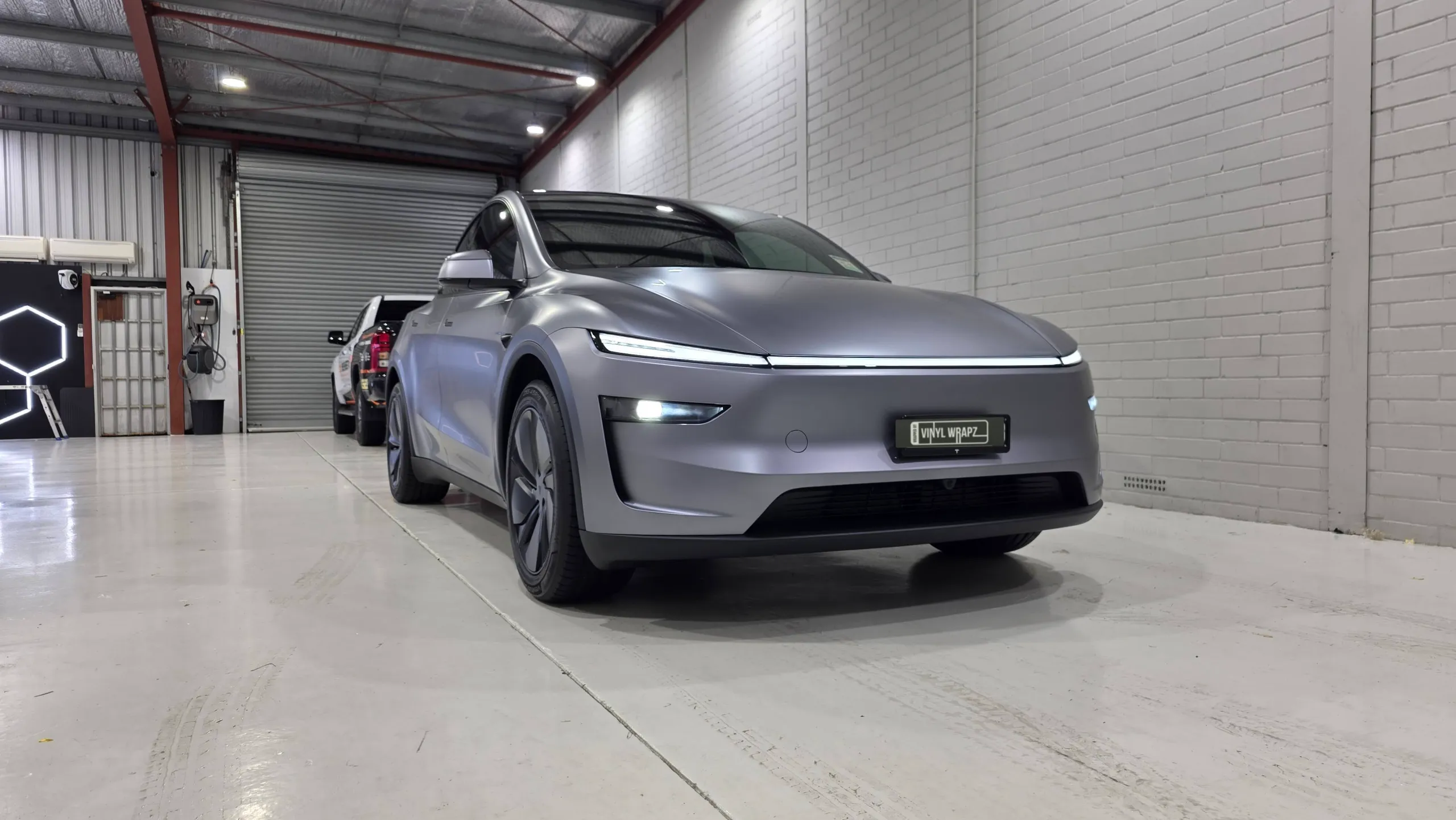 Satin grey Tesla Model Y with its front lights illuminated in the workshop