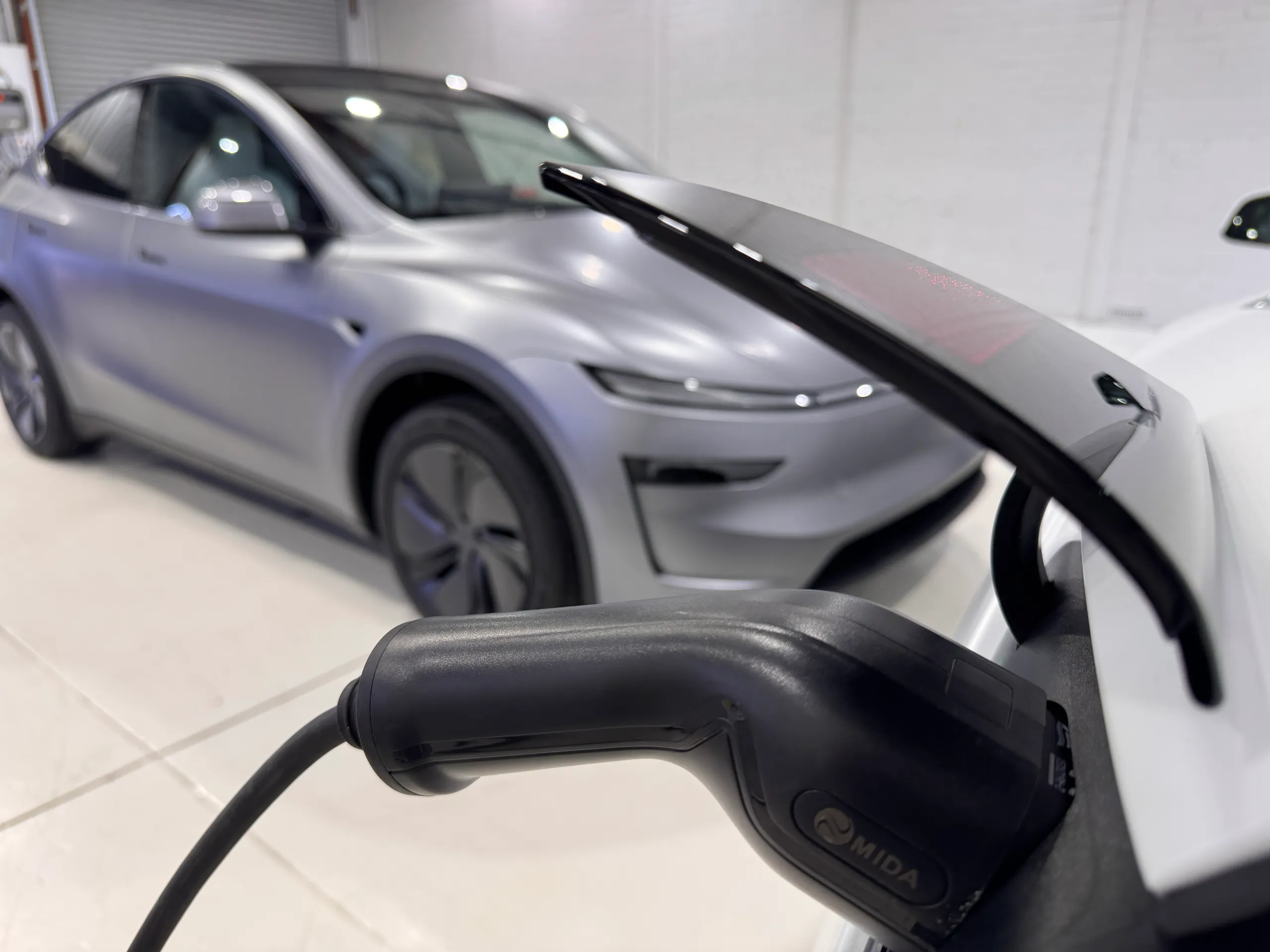 Charging connector plugged into a white car, with the satin grey Tesla Model Y behind it