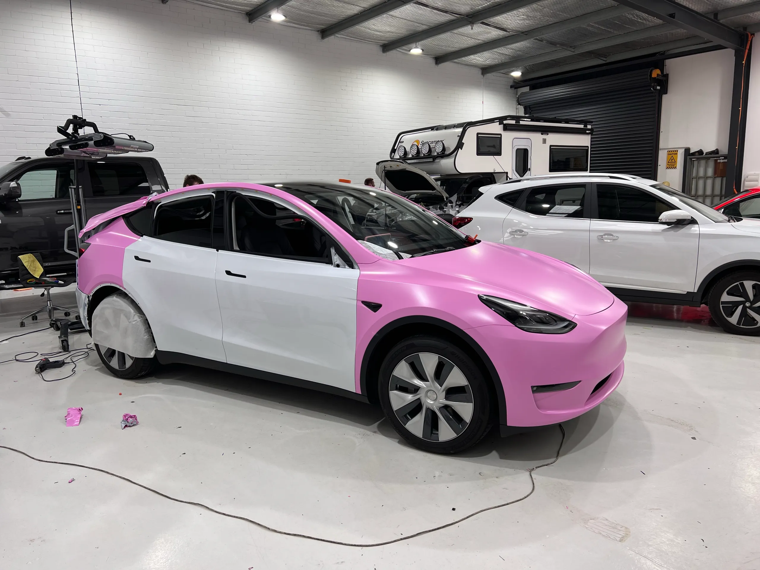 Tesla Model Y during a pink colour change, with white doors still visible and people in the workshop background