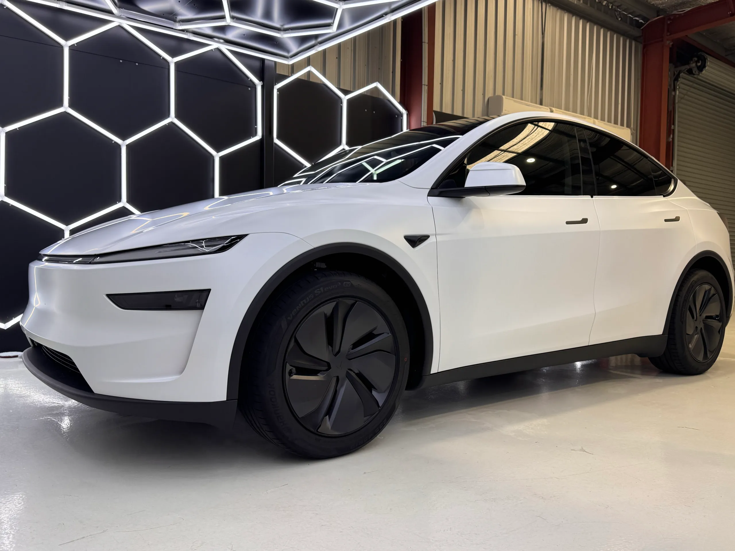 Matte white Tesla Model Y viewed from the front quarter beneath workshop lights