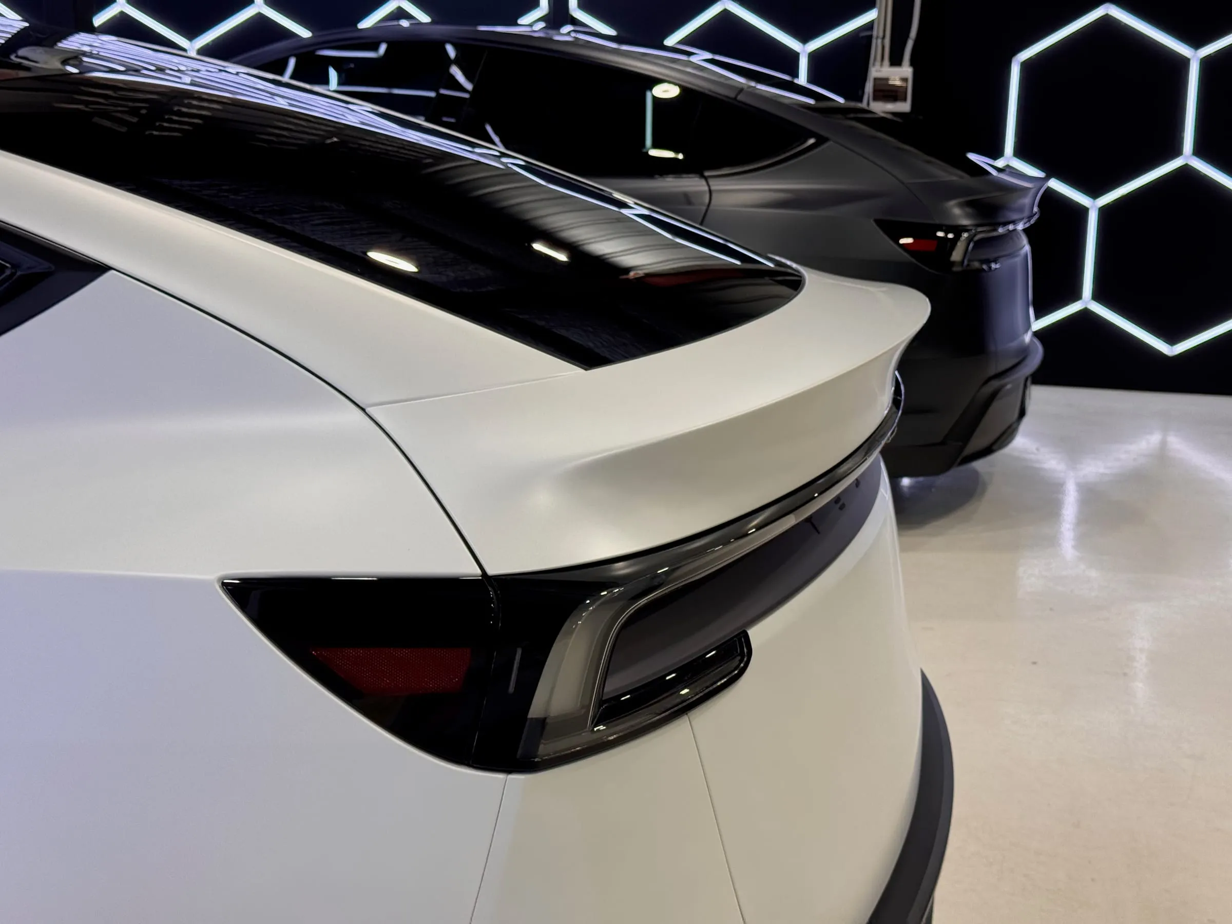 The rear spoiler edge and taillight of a white Tesla Model Y in clear paint protection film, a satin black Model Y behind it
