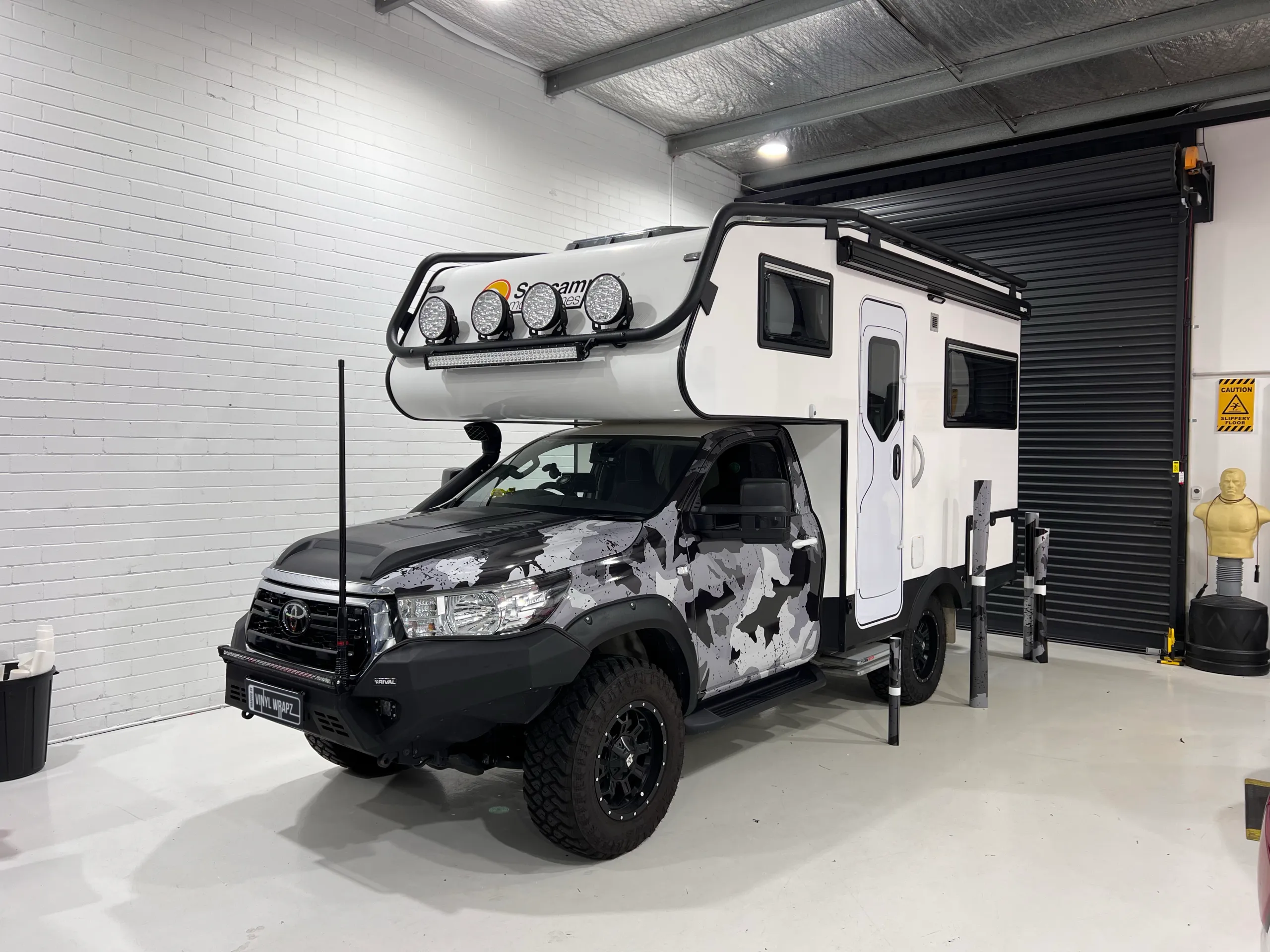 Camouflage-wrapped Hilux cab beneath a still-white Suncamper body during installation