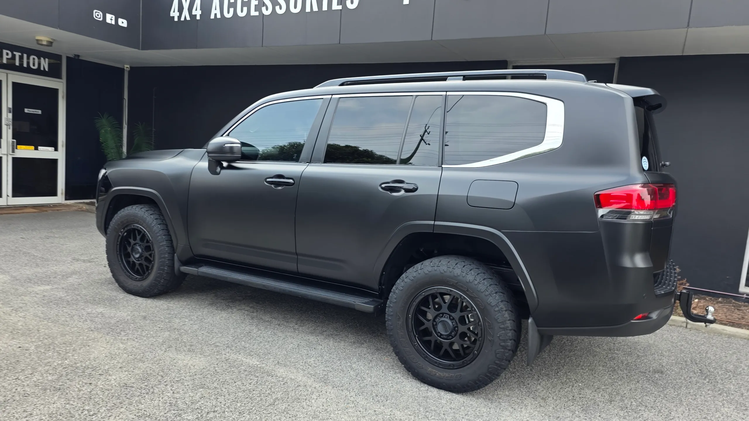 Satin black Toyota LandCruiser 300 viewed from the rear quarter outside an accessories shop