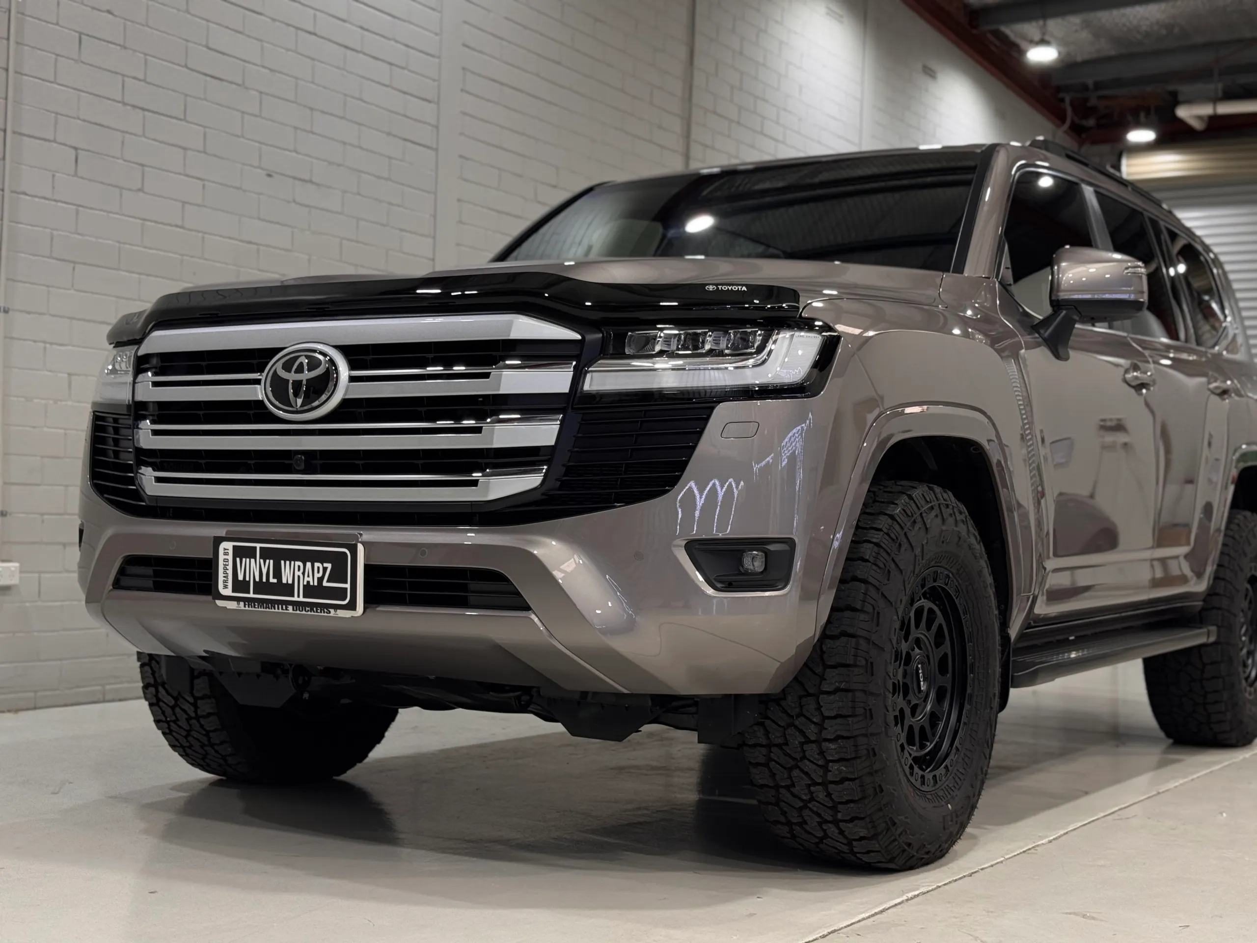 Low front-quarter view of the bronze-grey LandCruiser 300 grille, bonnet and black wheel