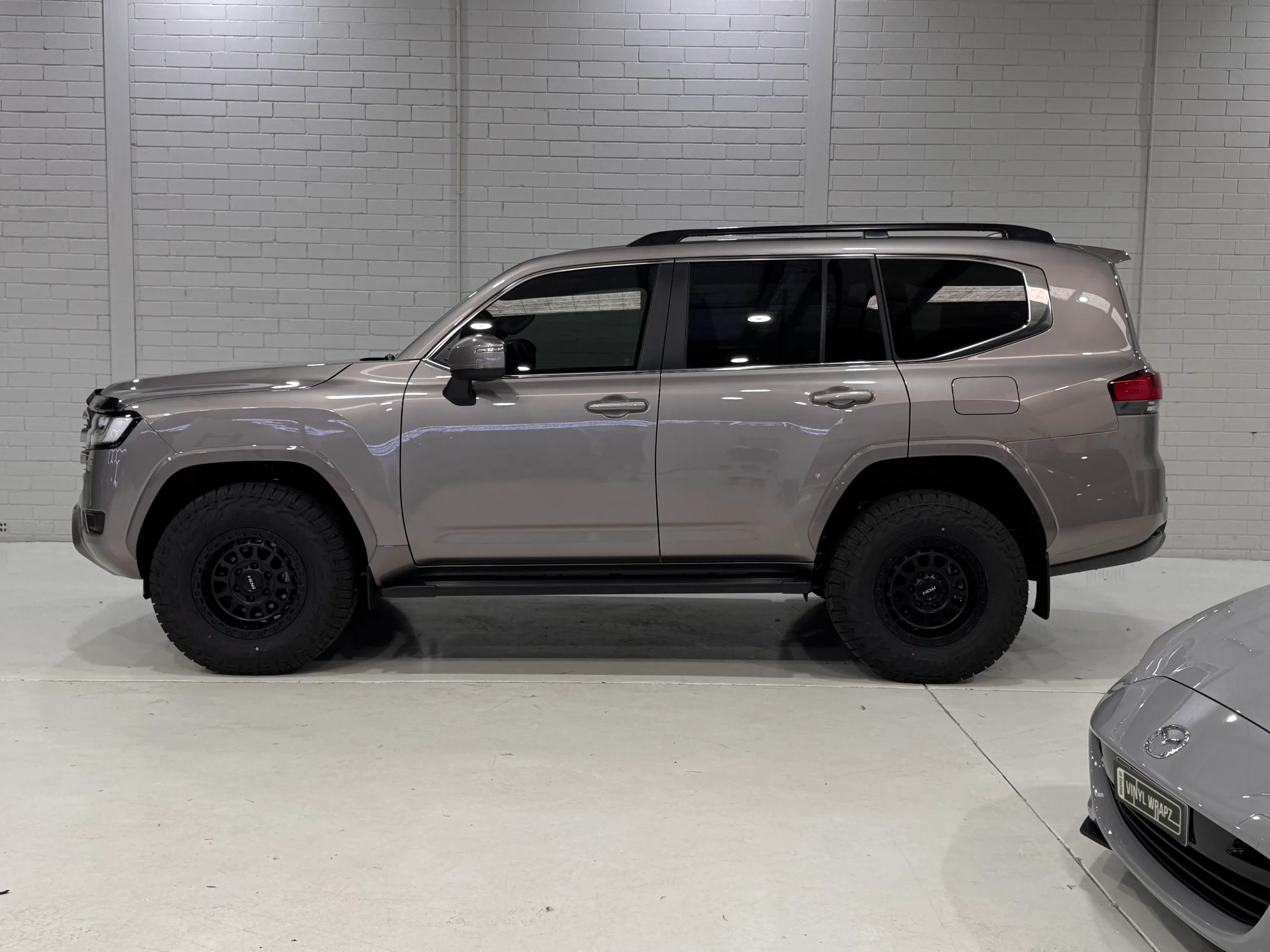 Side view of the bronze-grey LandCruiser 300 with black wheels and roof rails