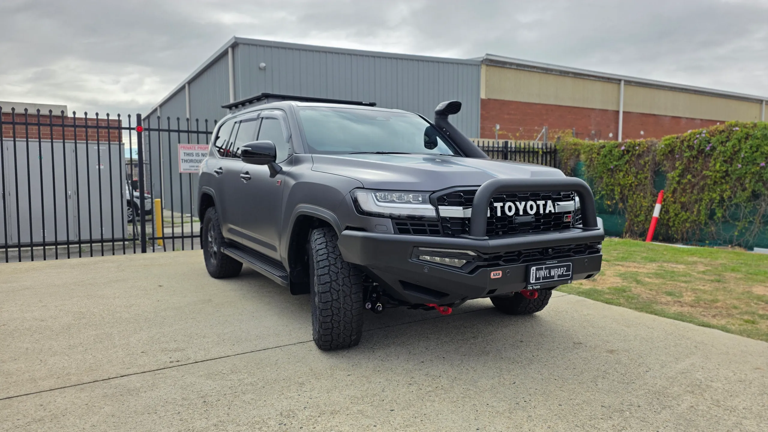 Matte grey LandCruiser 300 GR Sport with a bullbar and snorkel parked beside a fence