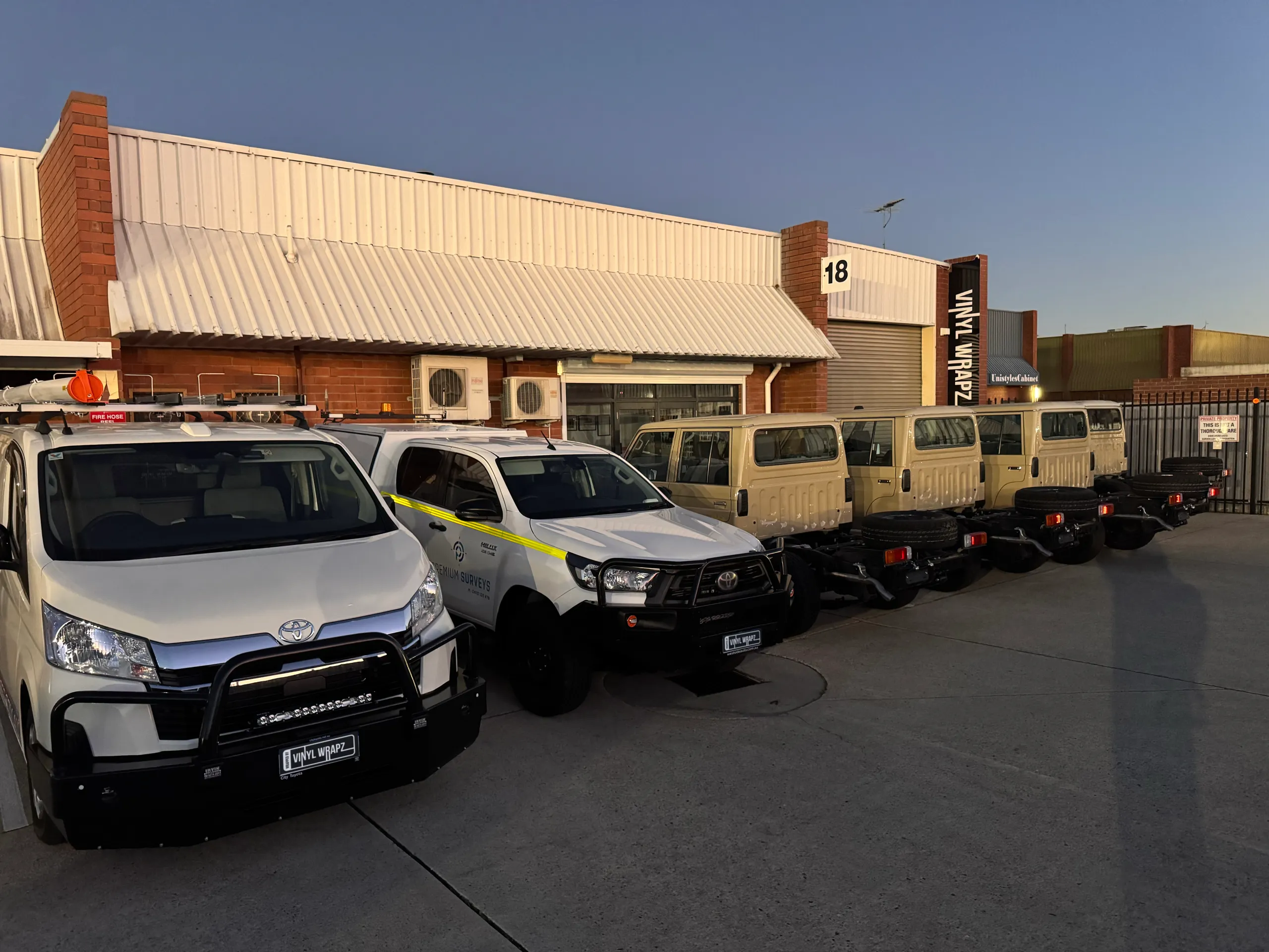 Tan LandCruiser cab-chassis vehicles lined up behind two white utes outside the workshop