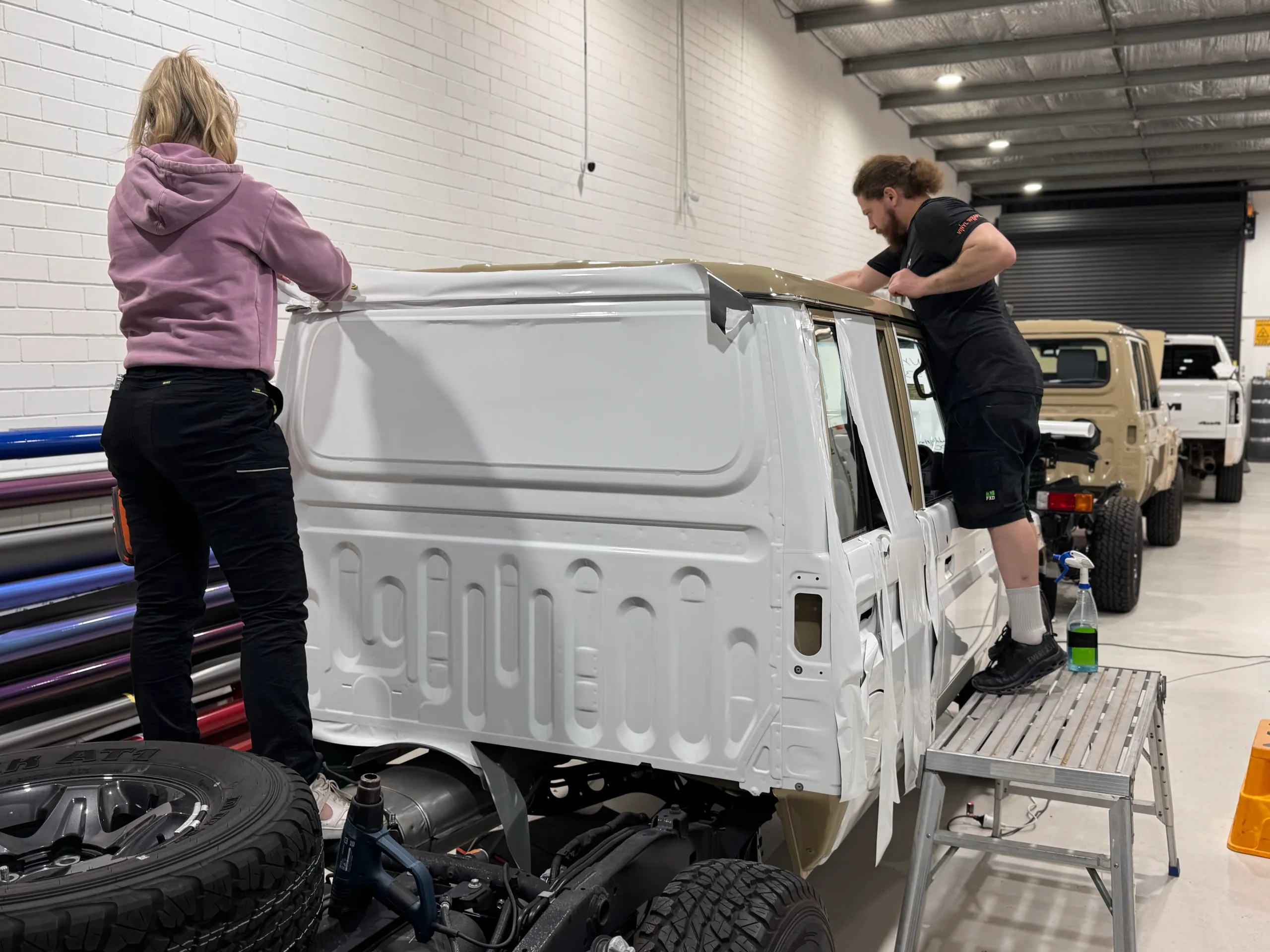 Installers fitting white film across the rear of a LandCruiser 79 cab above its exposed chassis