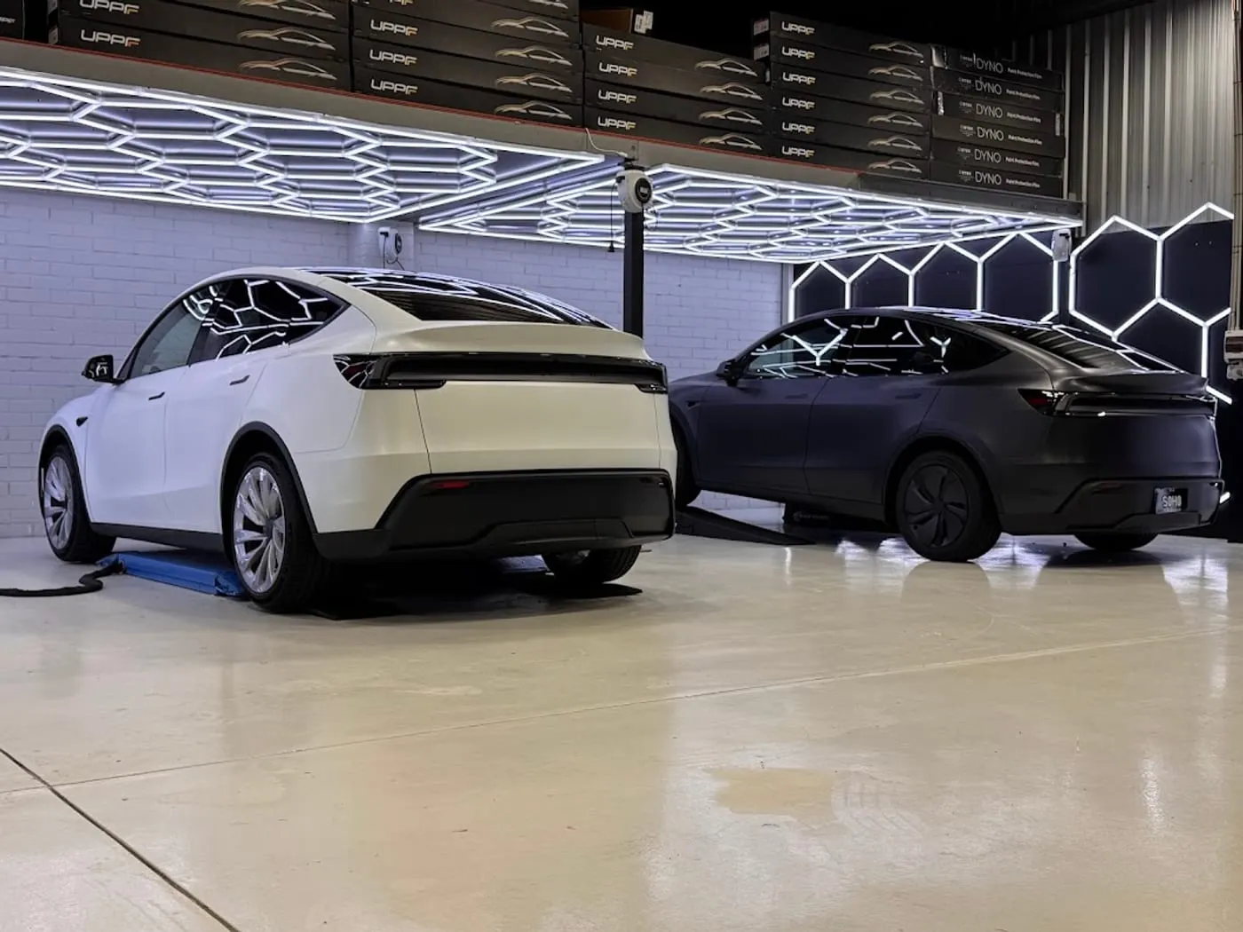 Two Tesla Model Y, one satin white and one satin black, nose to tail under the hexagon inspection lighting in the Vinyl Wrapz paint protection film bay, Osborne Park