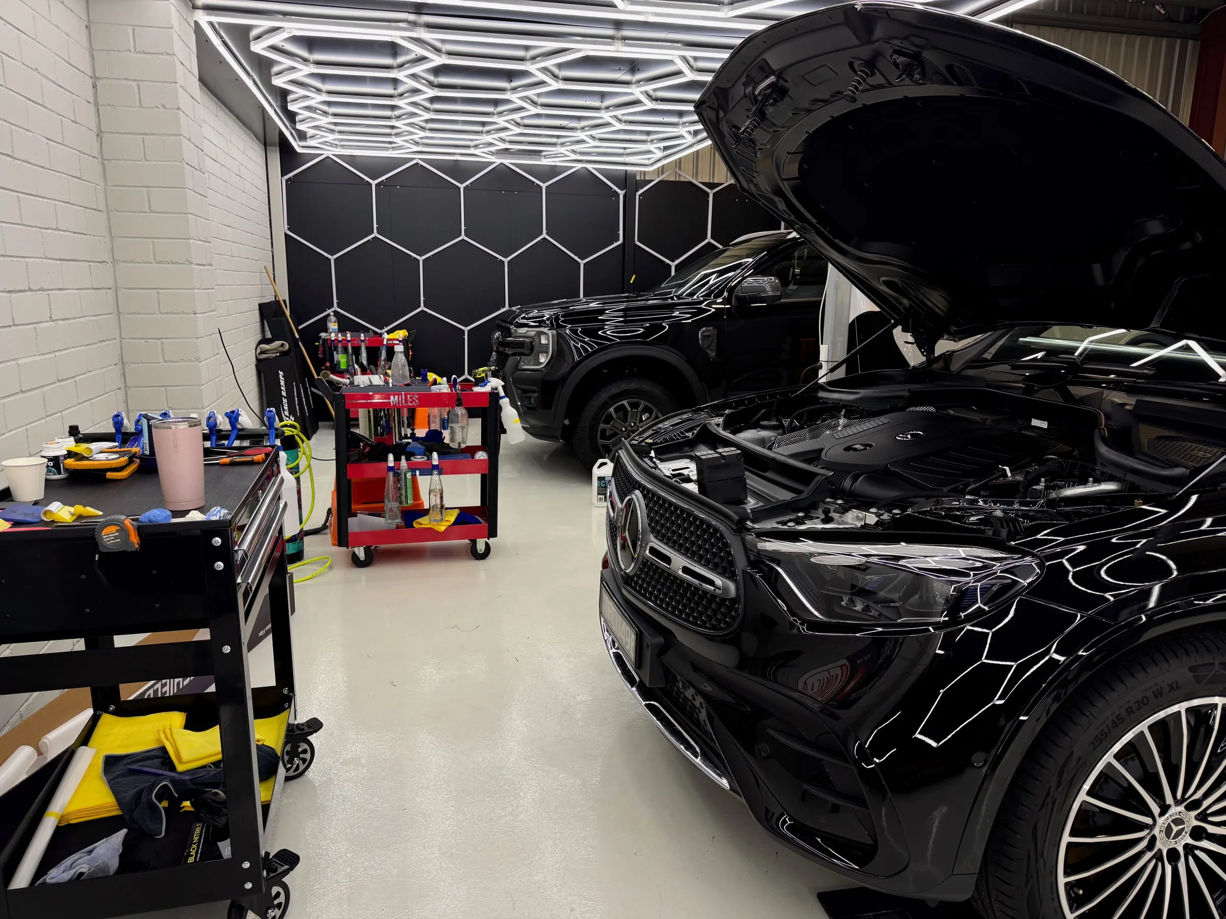 Black Mercedes GLE with its bonnet raised beside a Ford Ranger in the Vinyl Wrapz PPF workshop, tool carts alongside, under the hexagon inspection lighting