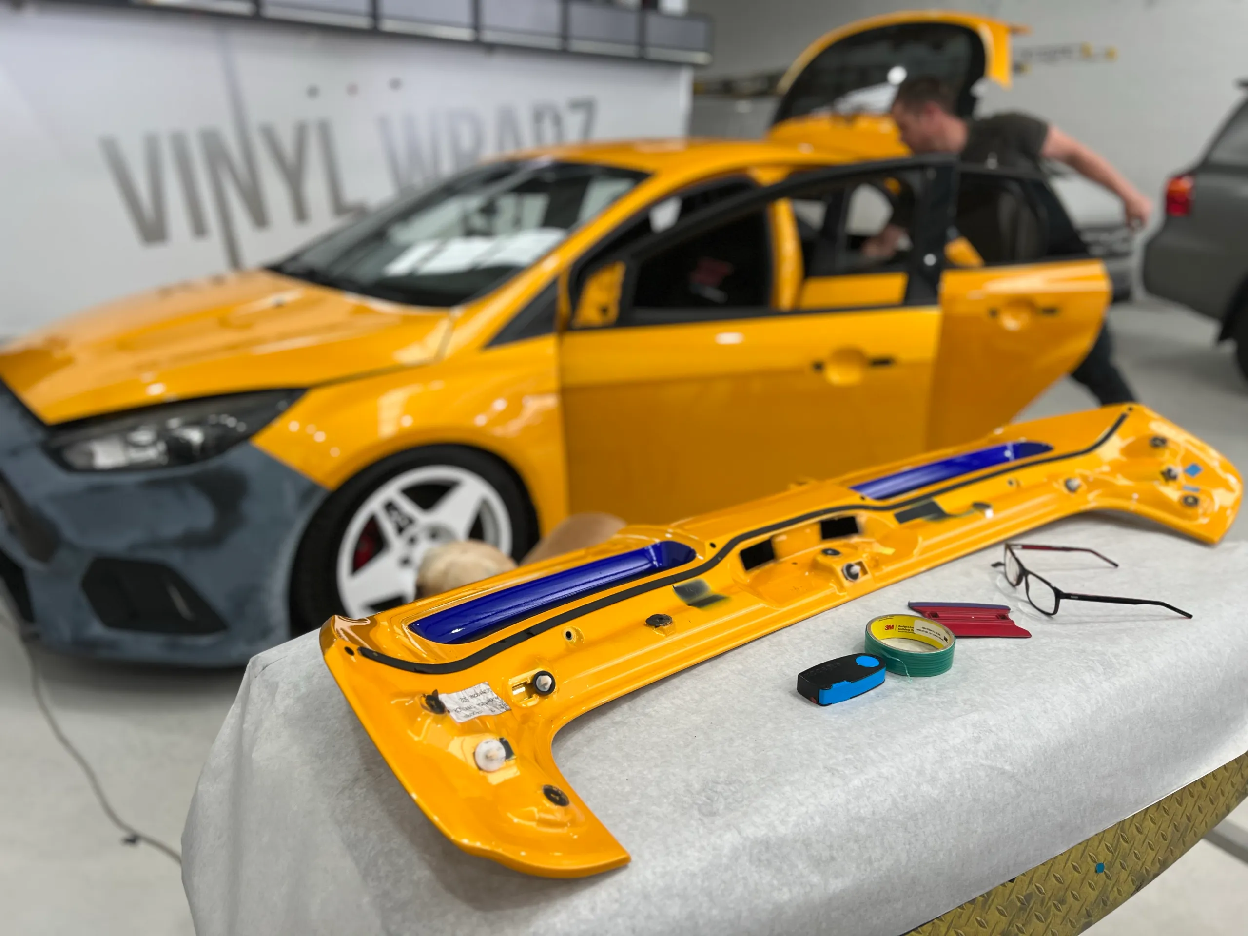 Yellow hatchback door removed for wrapping with an installer working beside the car